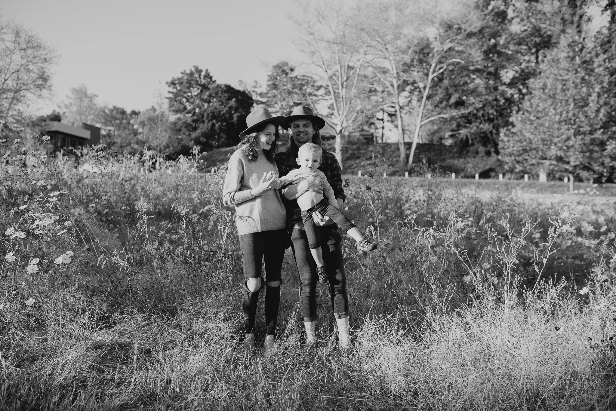 A black and white photo of a family of three standing outdoors in a field, with trees and a house in the background. The mother and father are holding their young child, and all are smiling or looking content. The family is dressed casually, with the