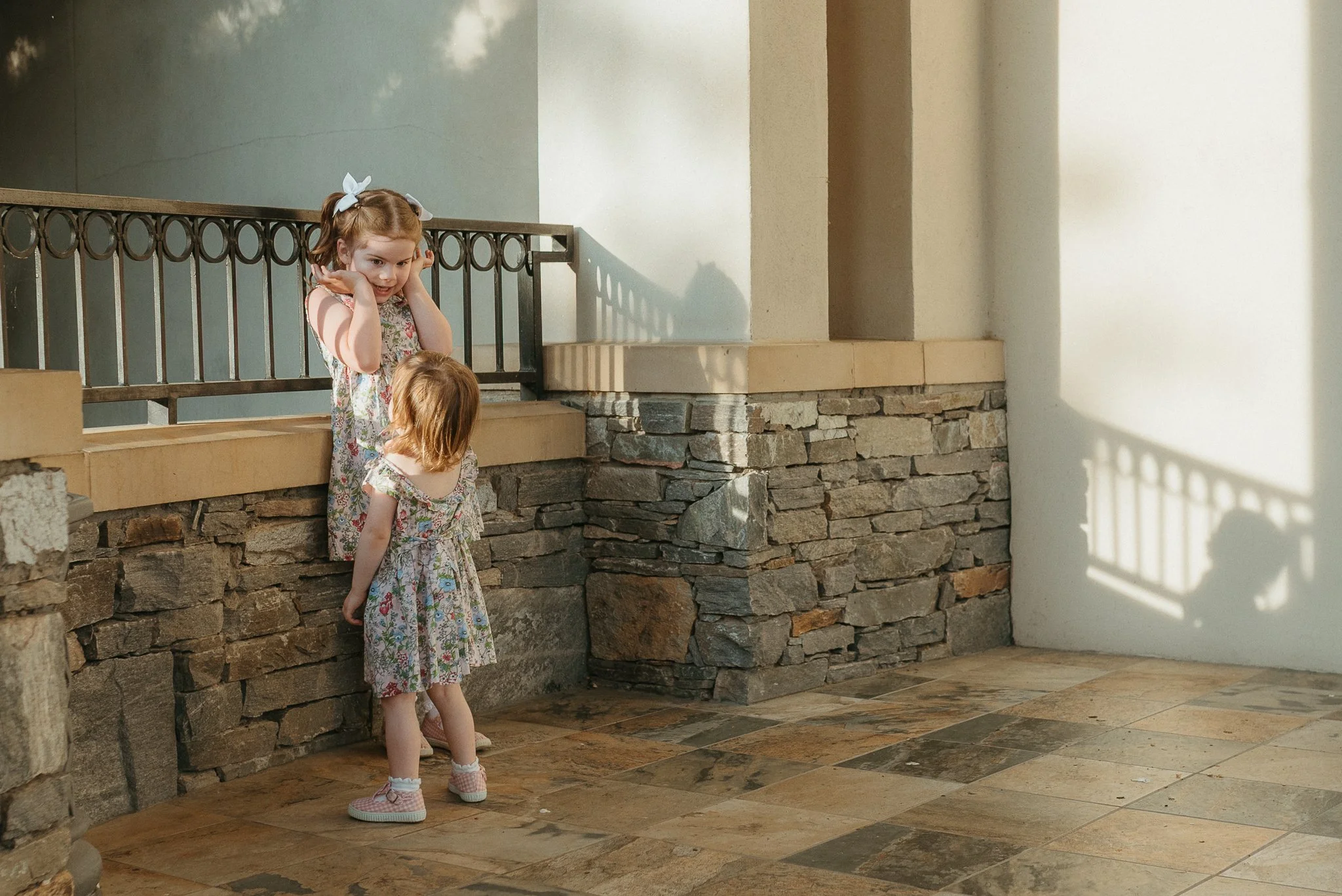 Two young girls with red hair, wearing floral dresses and pink shoes, standing near a stone and metal railing inside a building with stone and white walls, and sunlight casting shadows.