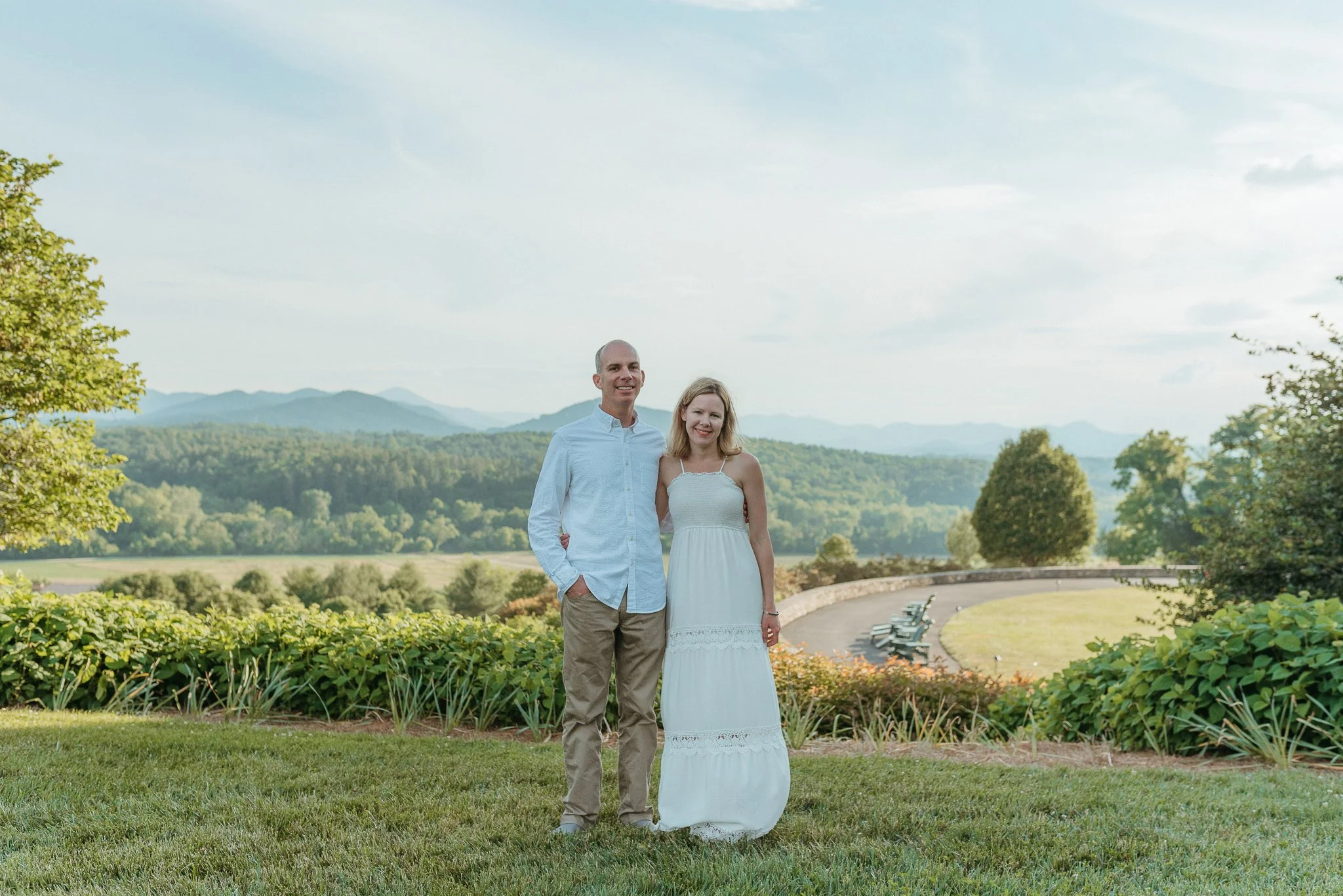 A couple standing in a park with greenery and mountains in the background, smiling at the camera.
