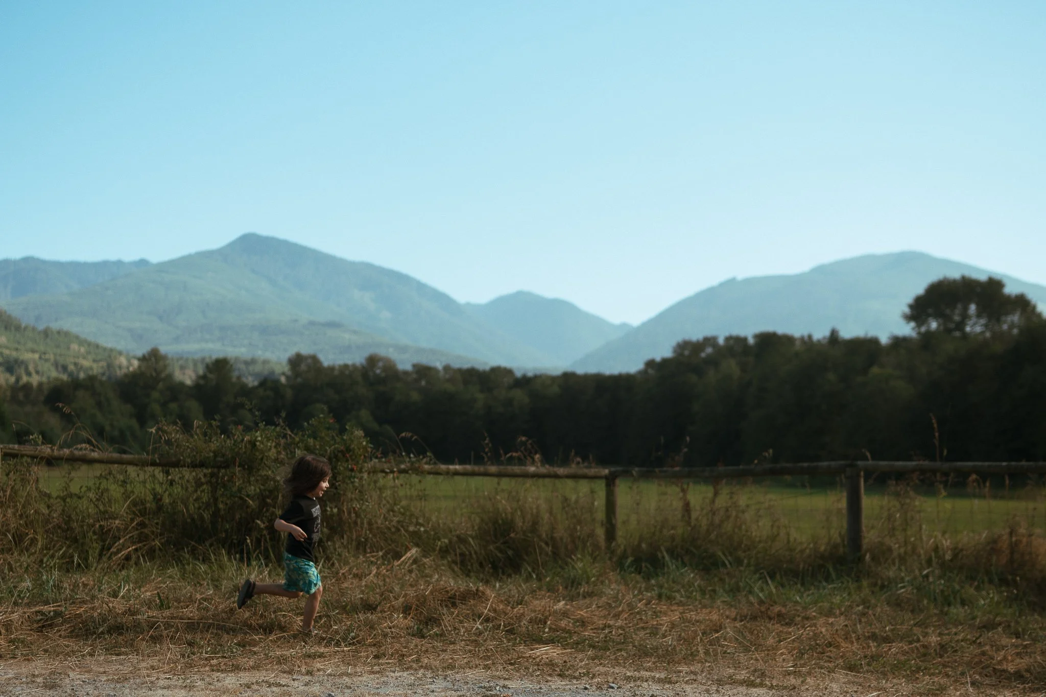 A young girl running outdoors in front of a wooden fence with hills and mountains in the background under a clear blue sky.