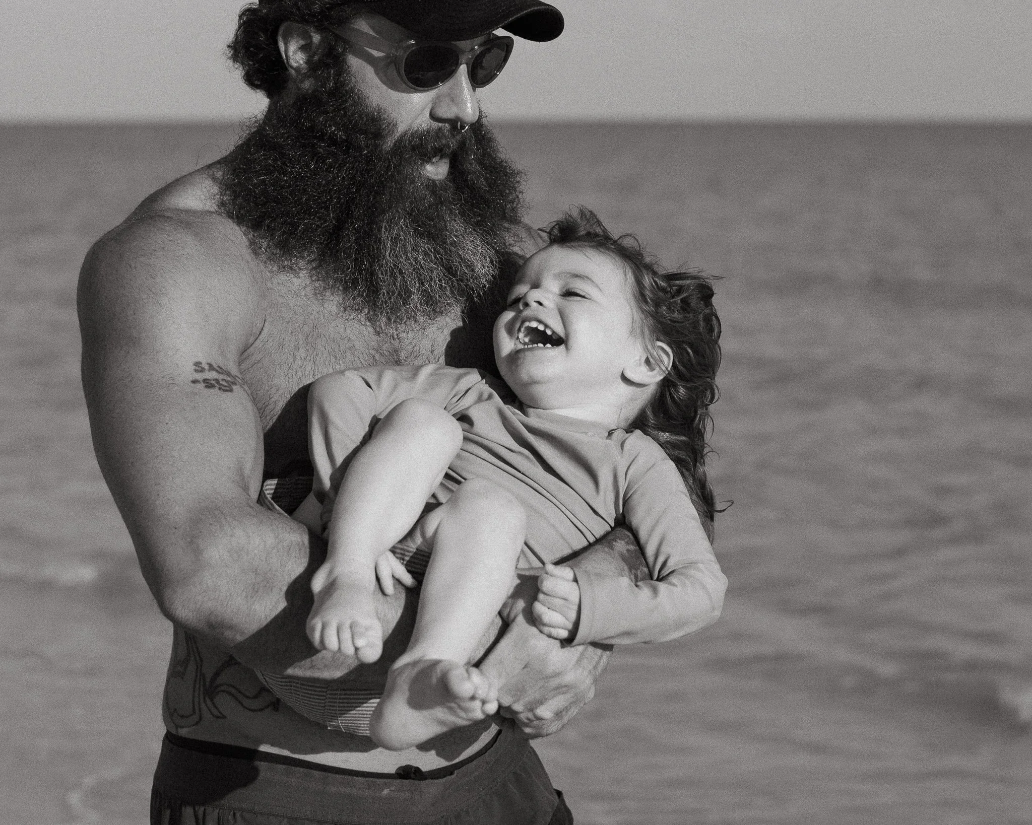 A man with a thick beard, wearing sunglasses and a cap, is holding a young girl. The girl is smiling with her head tilted back and eyes closed, showing her teeth, in an outdoor setting with a calm body of water in the background.