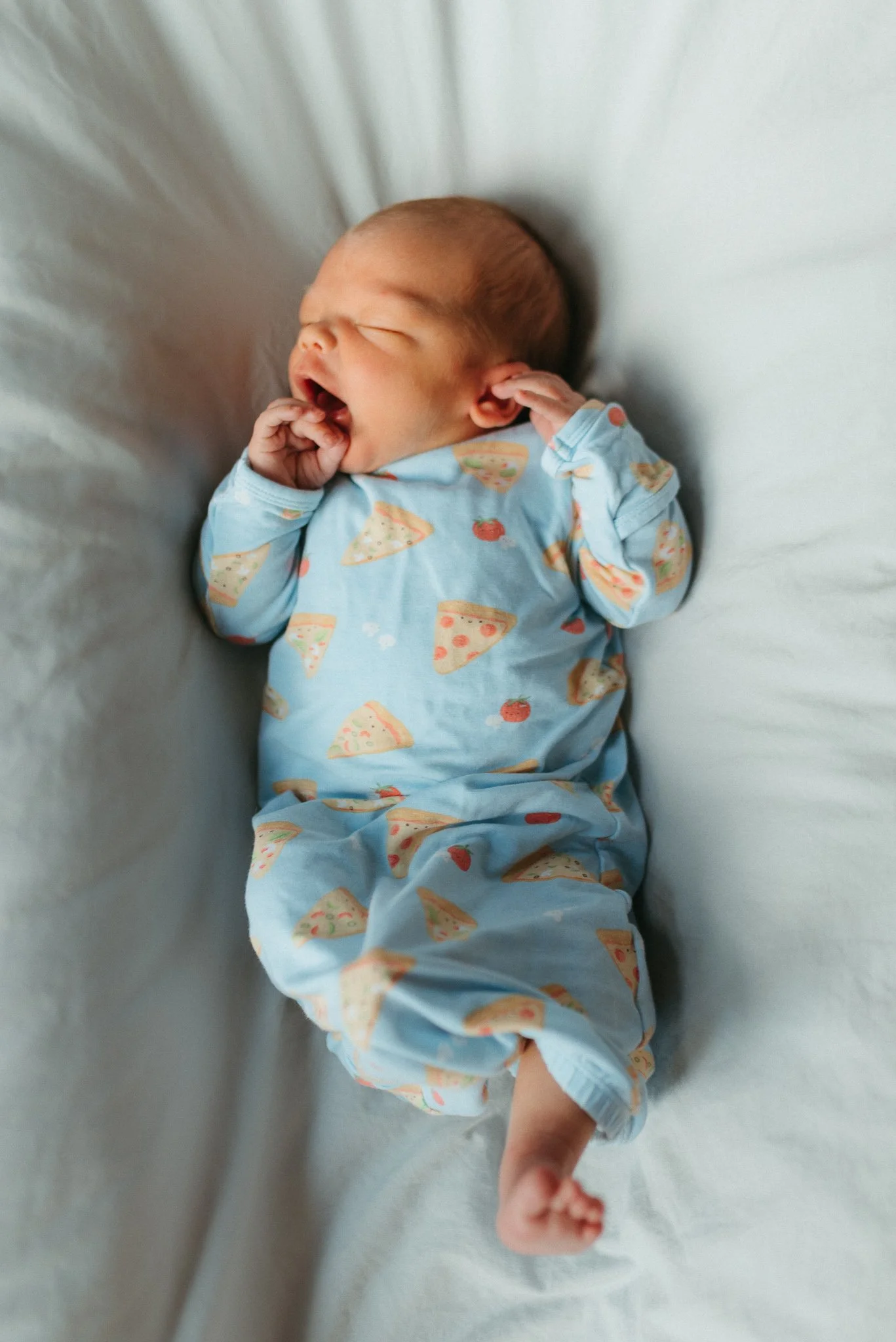 Newborn baby sleeping on a white bed, wearing a blue pajama with pizza and tomato patterns, with arms bent and hands near face.
