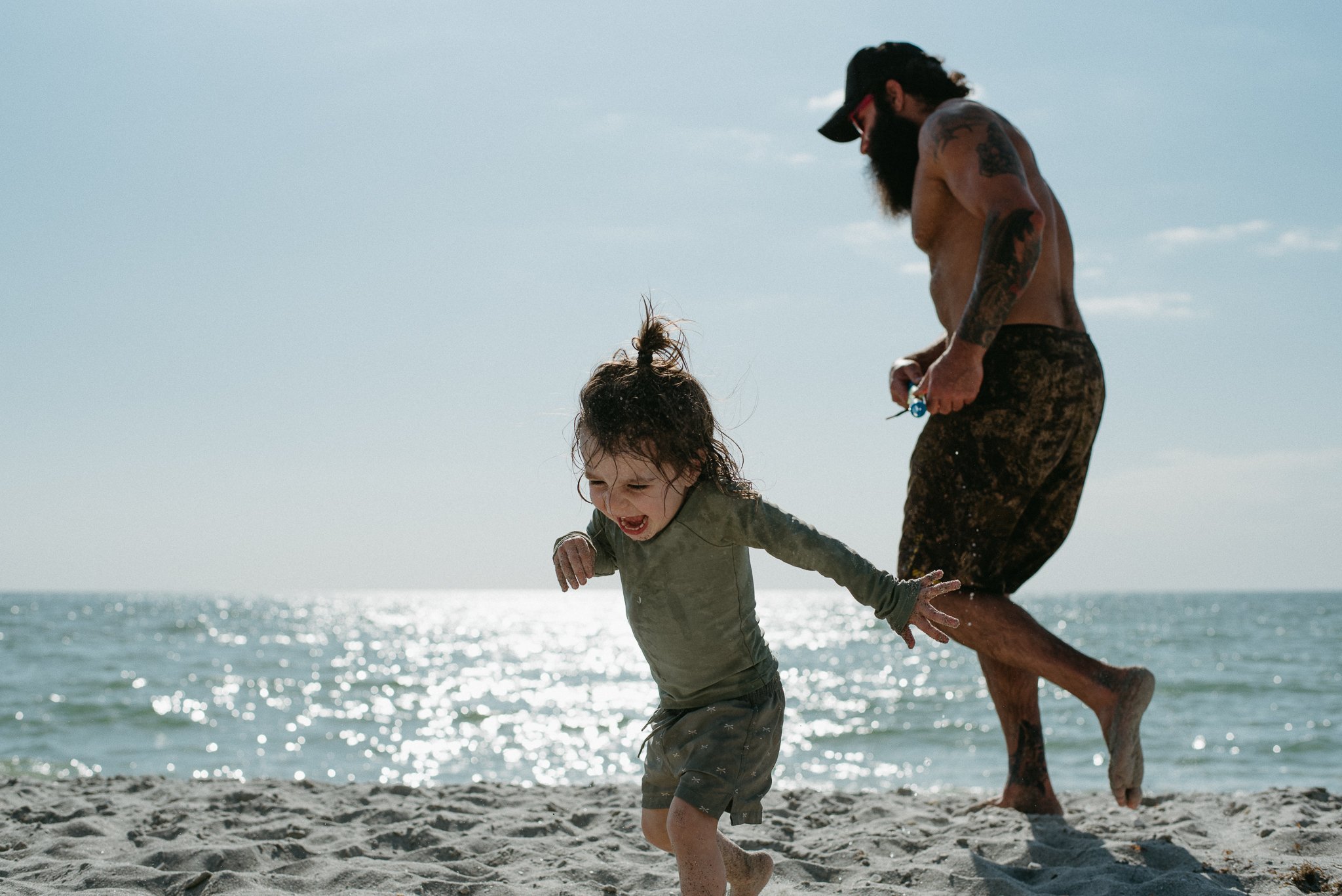 A bearded man with tattoos, wearing sunglasses and camouflage shorts, is holding a young girl’s hands as they play and run on the beach near the water, with the ocean and sky in the background.