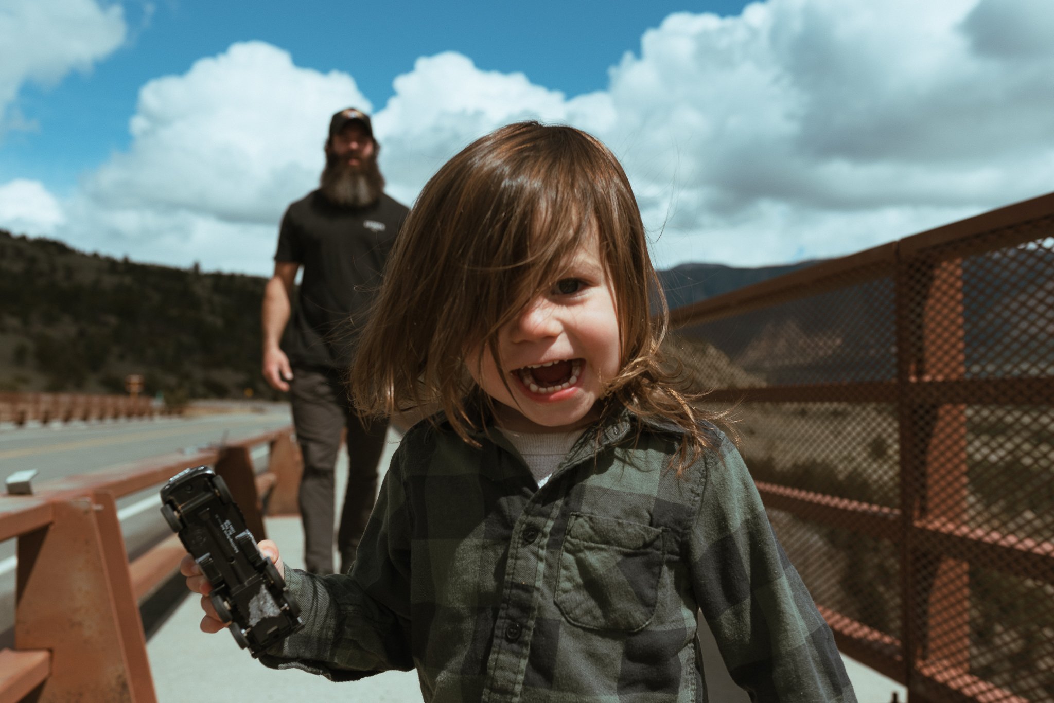 A young girl with long, curly hair laughing, holding a flashlight, on a bridge with a man in the background, mountainous landscape and blue sky with clouds.