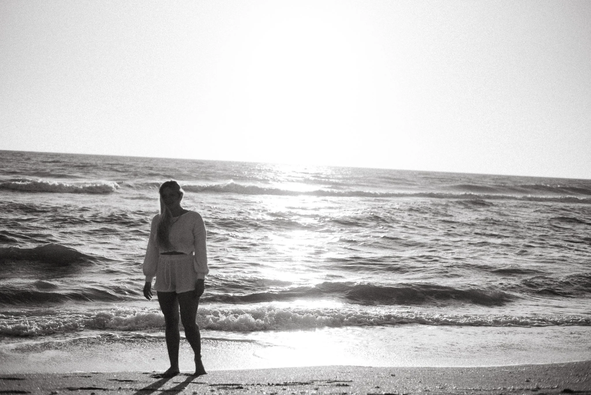 A woman stands on the beach at sunset, facing the ocean, with waves approaching her feet.