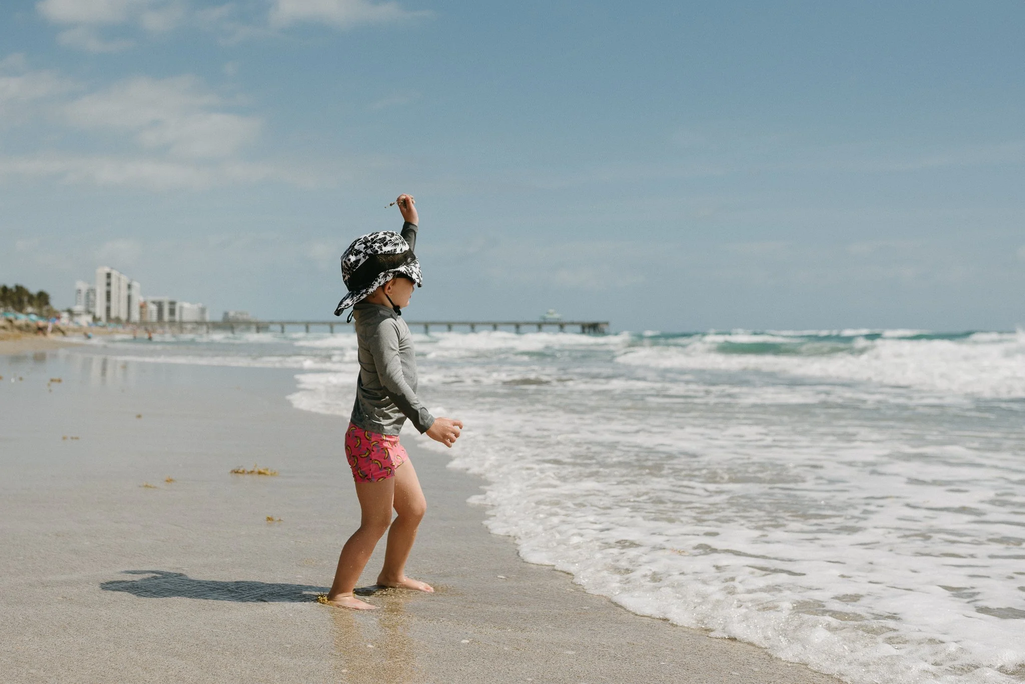 Child in pink shorts and a hat walking on the beach near the water, with buildings and a pier in the background.
