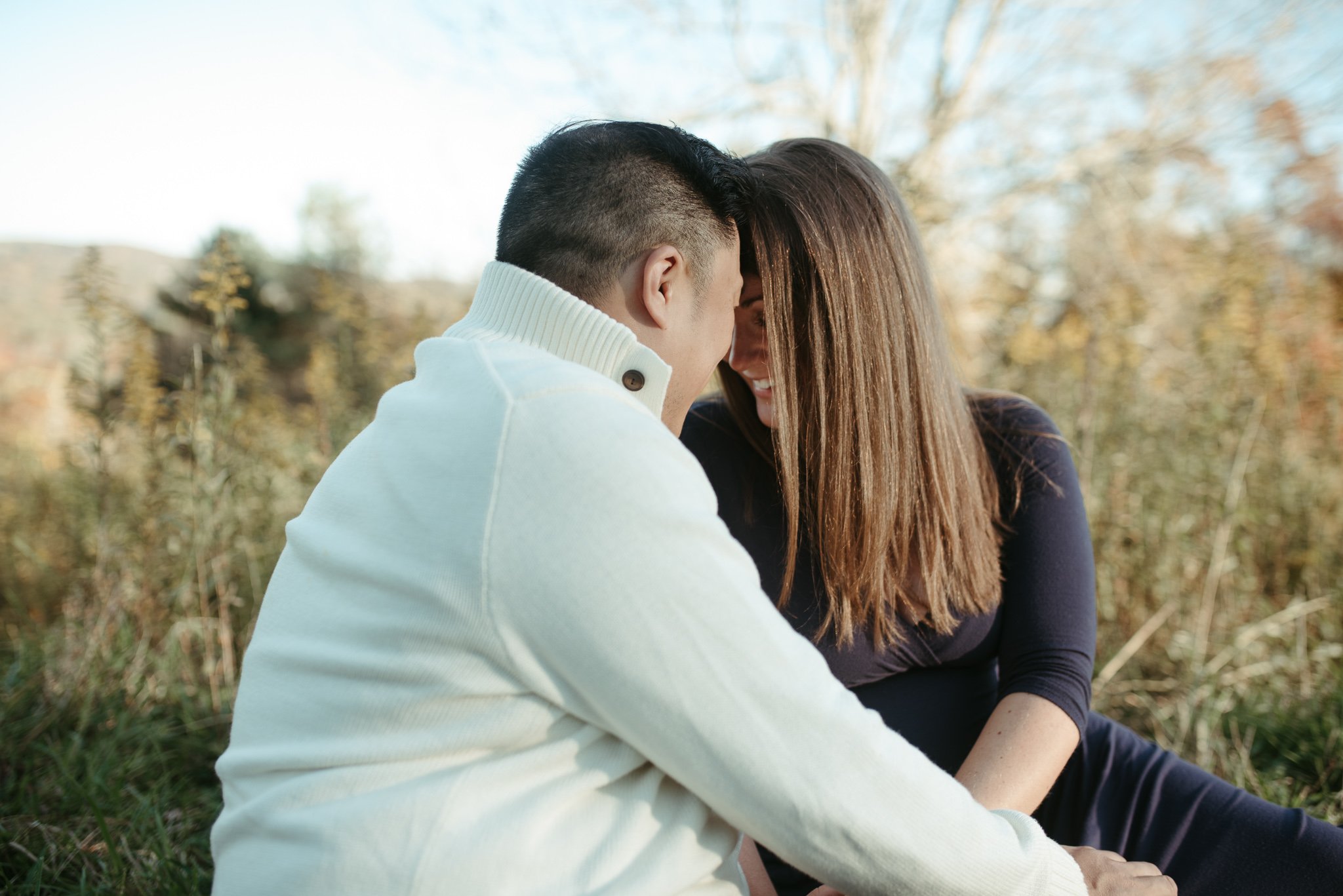 A couple sitting closely outdoors during autumn, with trees in the background, smiling and leaning their foreheads together.