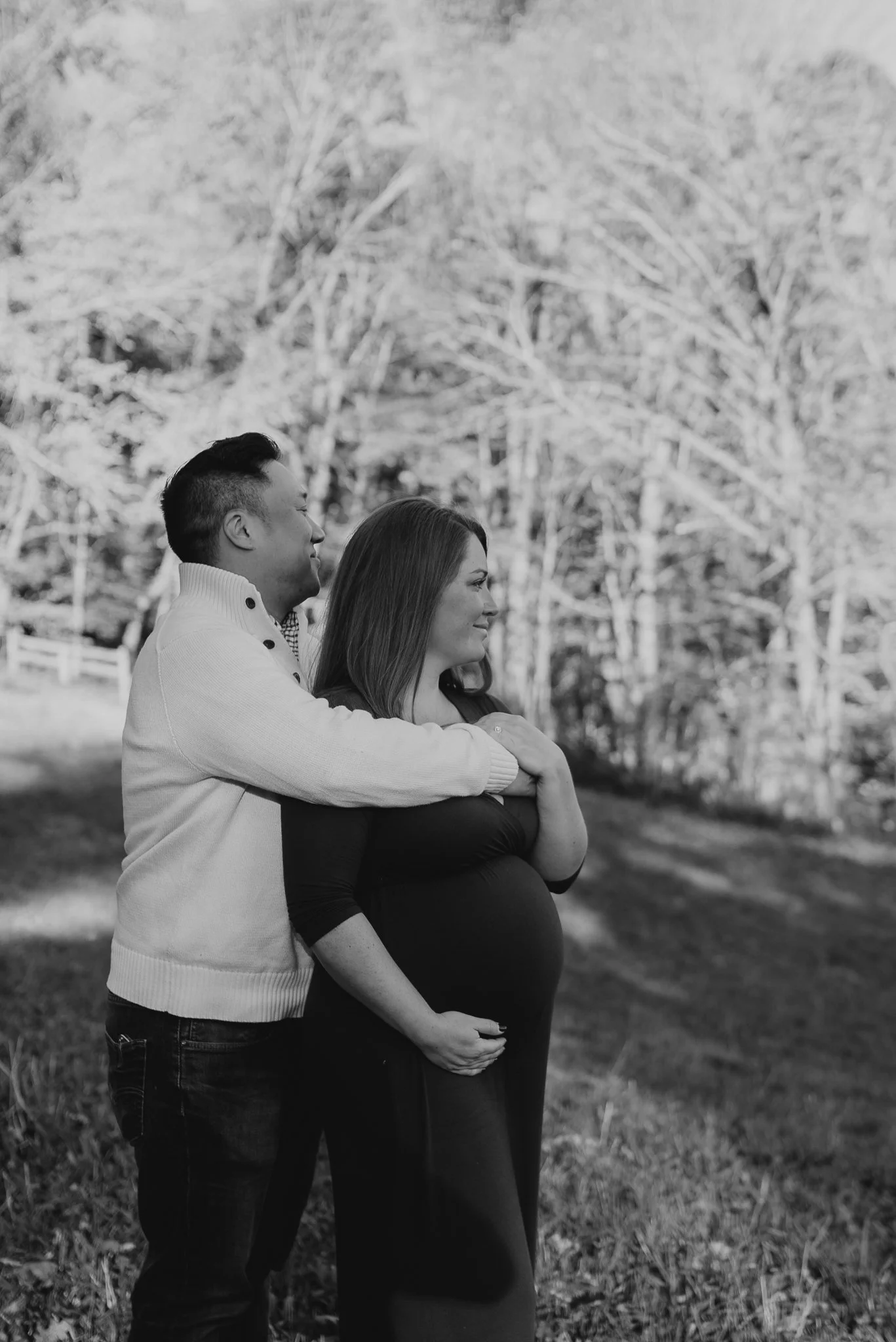 A couple, with the man embracing the pregnant woman from behind, stands outside with trees in the background. The image is in black and white.