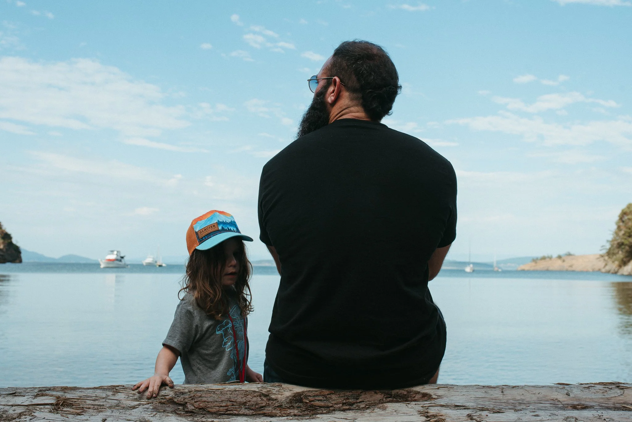 A man with a beard and glasses faces away as he stands by a body of water, with a young girl in a hat and gray shirt beside him, near a fallen log, with boats and mountains in the background under a partly cloudy sky.