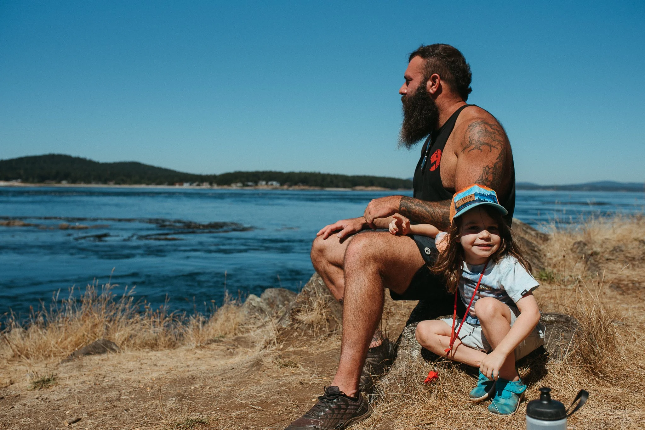 A man with a beard and tattoos sitting on a rock by the water with a young girl in front of him. The girl is wearing a cap and colorful shoes, smiling. The scene is outdoors near a lake or river, with mountains or hills in the background under a clea