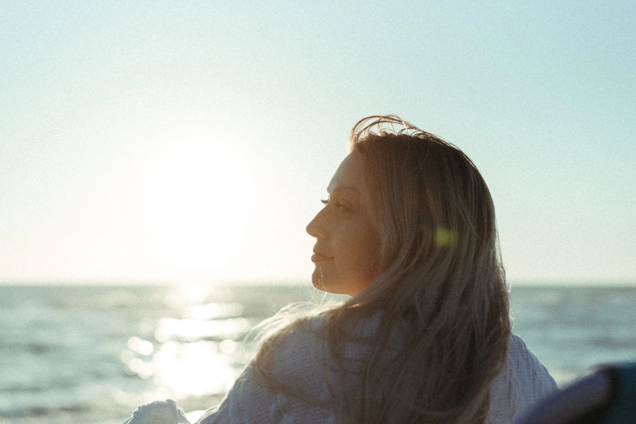 A woman with long dark hair sitting by the ocean at sunset, looking serene and contemplative.