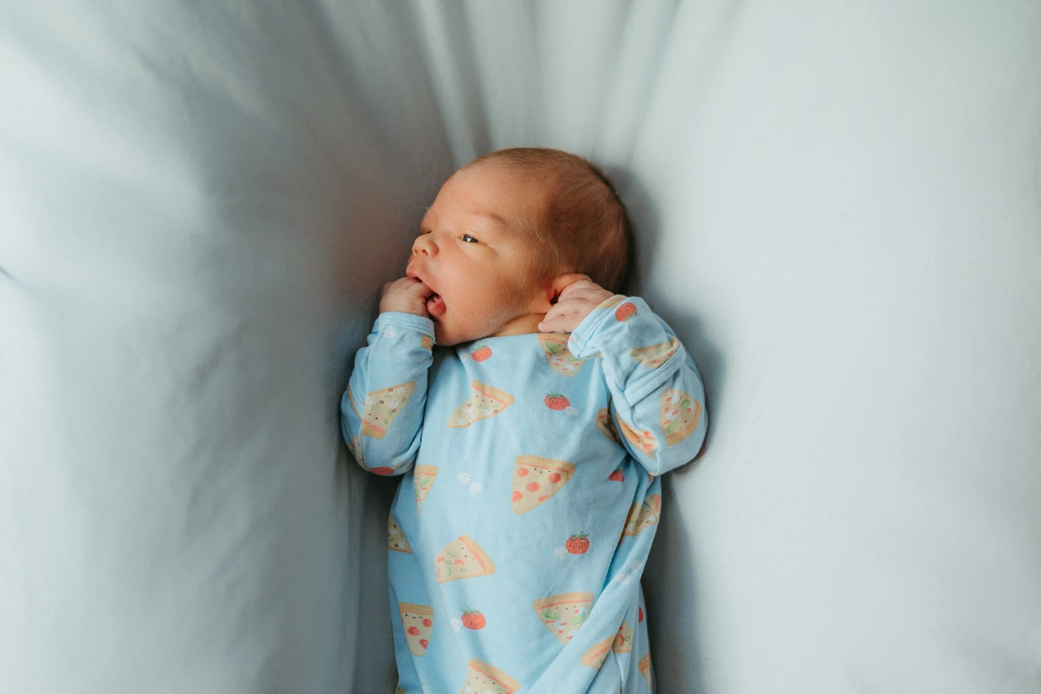 A baby lying on its side in a crib or bassinet, wearing a blue onesie with ice cream and strawberry patterns, with a puffed cheek, against a plain wall.