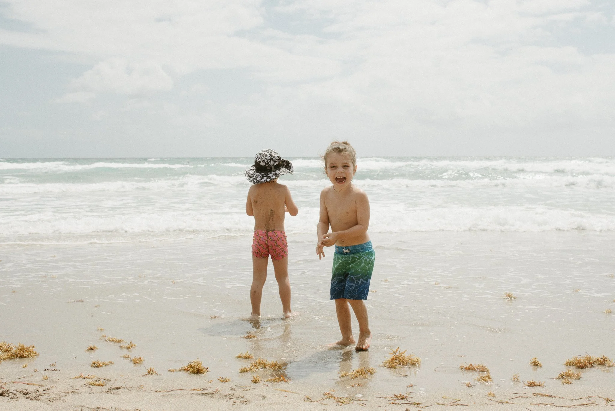 Two smiling children, a girl and a boy, playing at the beach with ocean waves in the background. The girl has blonde hair and the boy has dark hair with a hat, both wearing swim trunks.