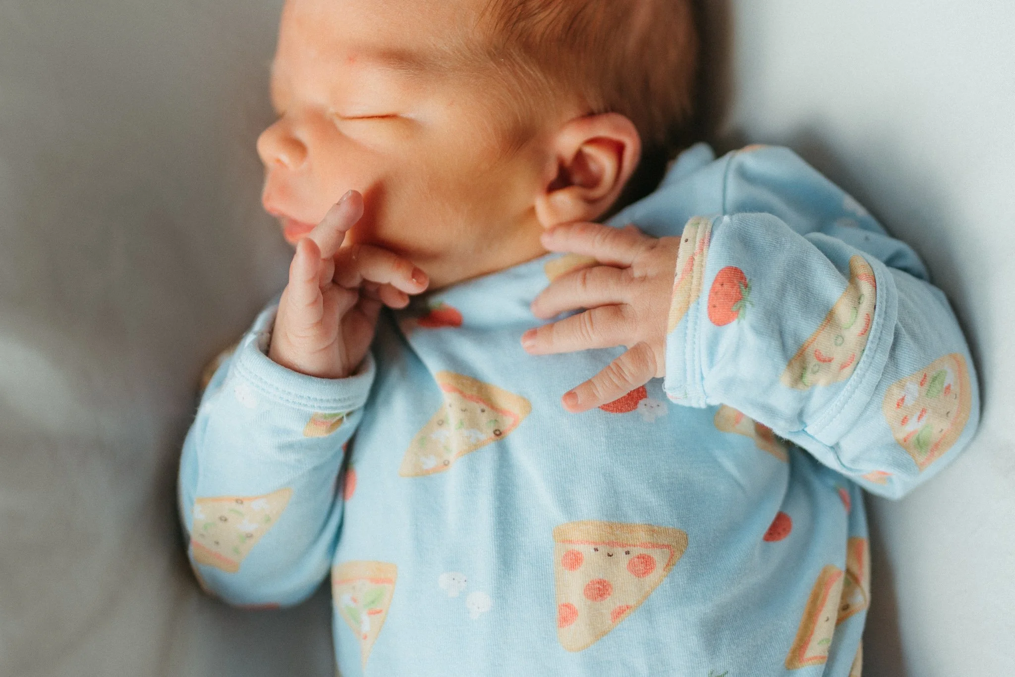 A sleeping baby with a pacifier and a hand near their face, wearing a blue shirt with pizza and strawberry patterns.