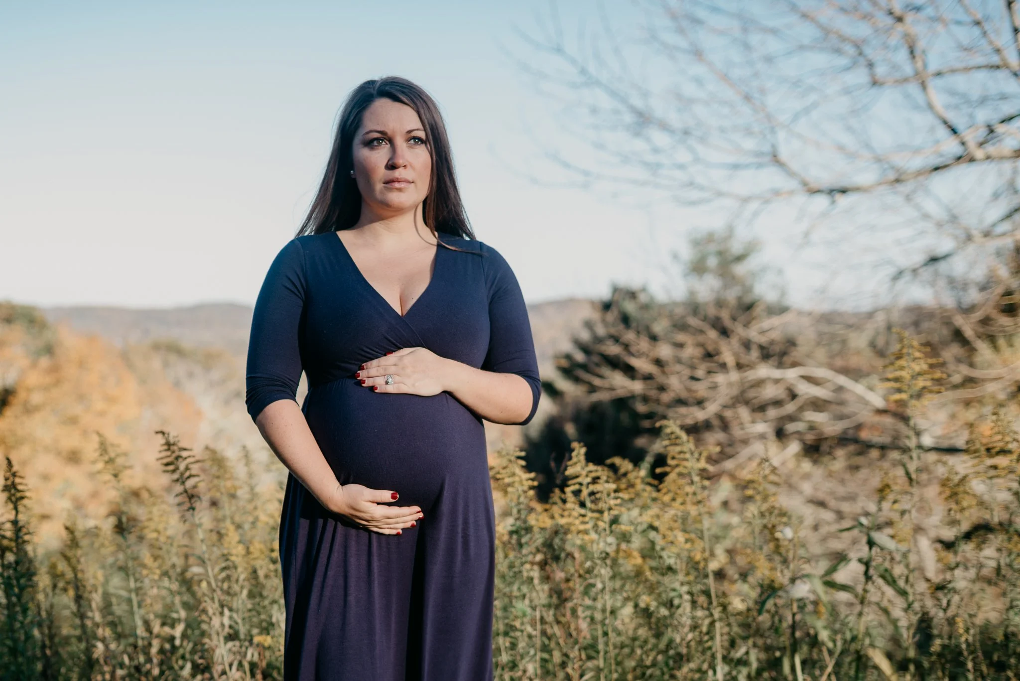 A pregnant woman in a dark blue dress standing outdoors near trees with autumn foliage, holding her baby bump with one hand and looking into the distance.