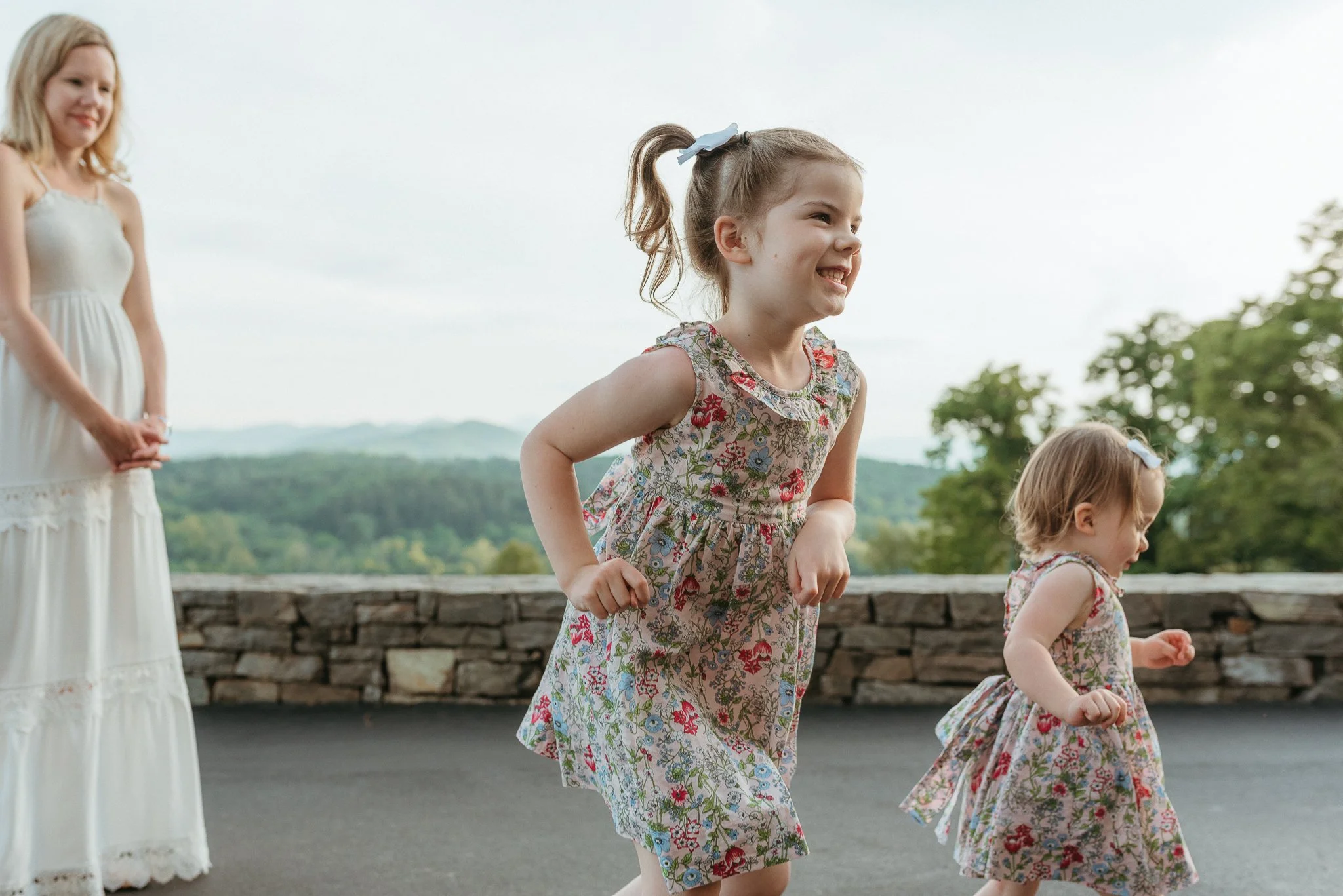 Two young girls, wearing matching floral dresses, are outdoors and appear to be playing or running. A woman in a white dress is standing nearby, watching them. The background shows a scenic view of distant mountains and greenery.