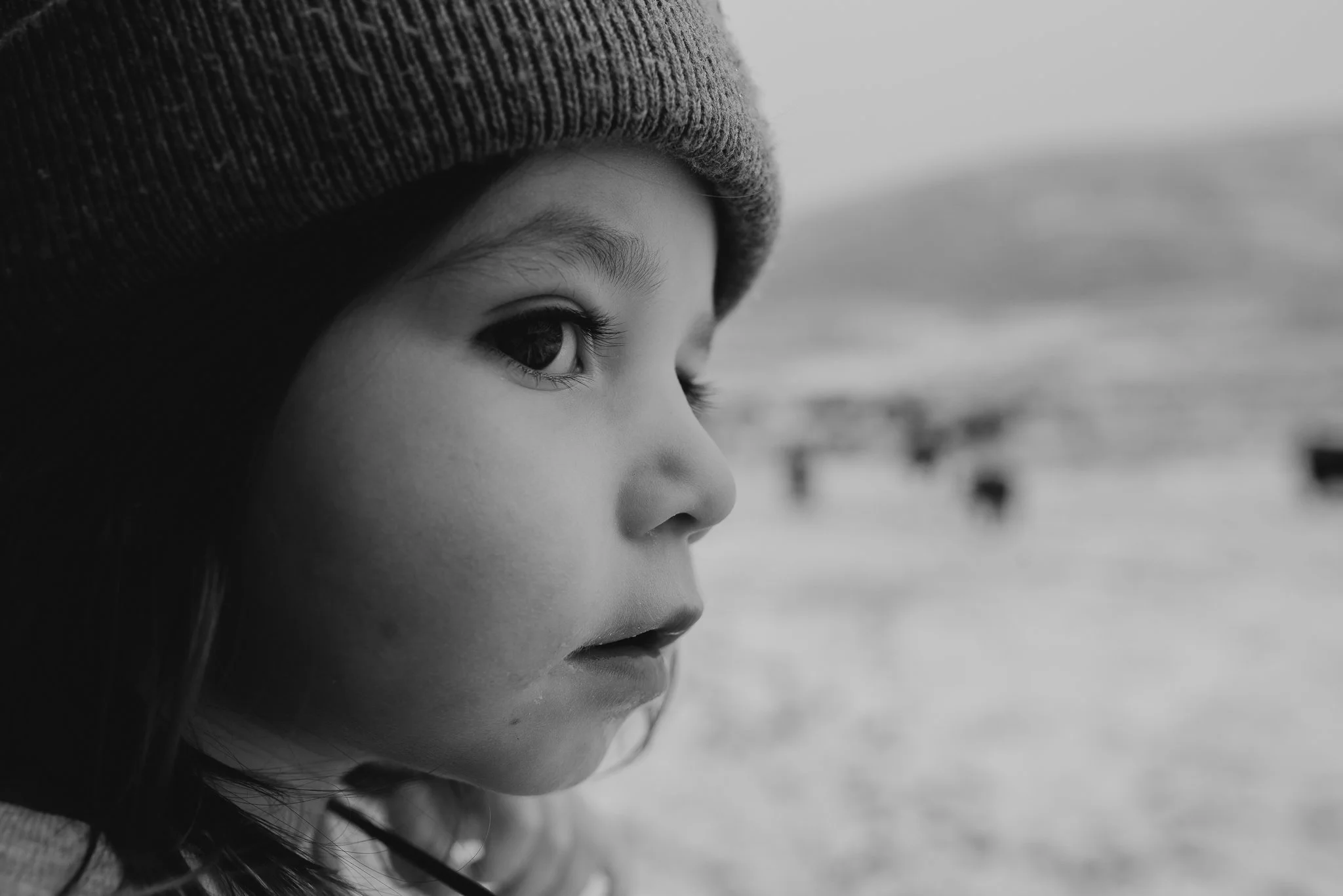 Close-up black-and-white photo of a young girl's face in profile, wearing a knit hat. She is looking towards a distant landscape with blurred structures in the background.