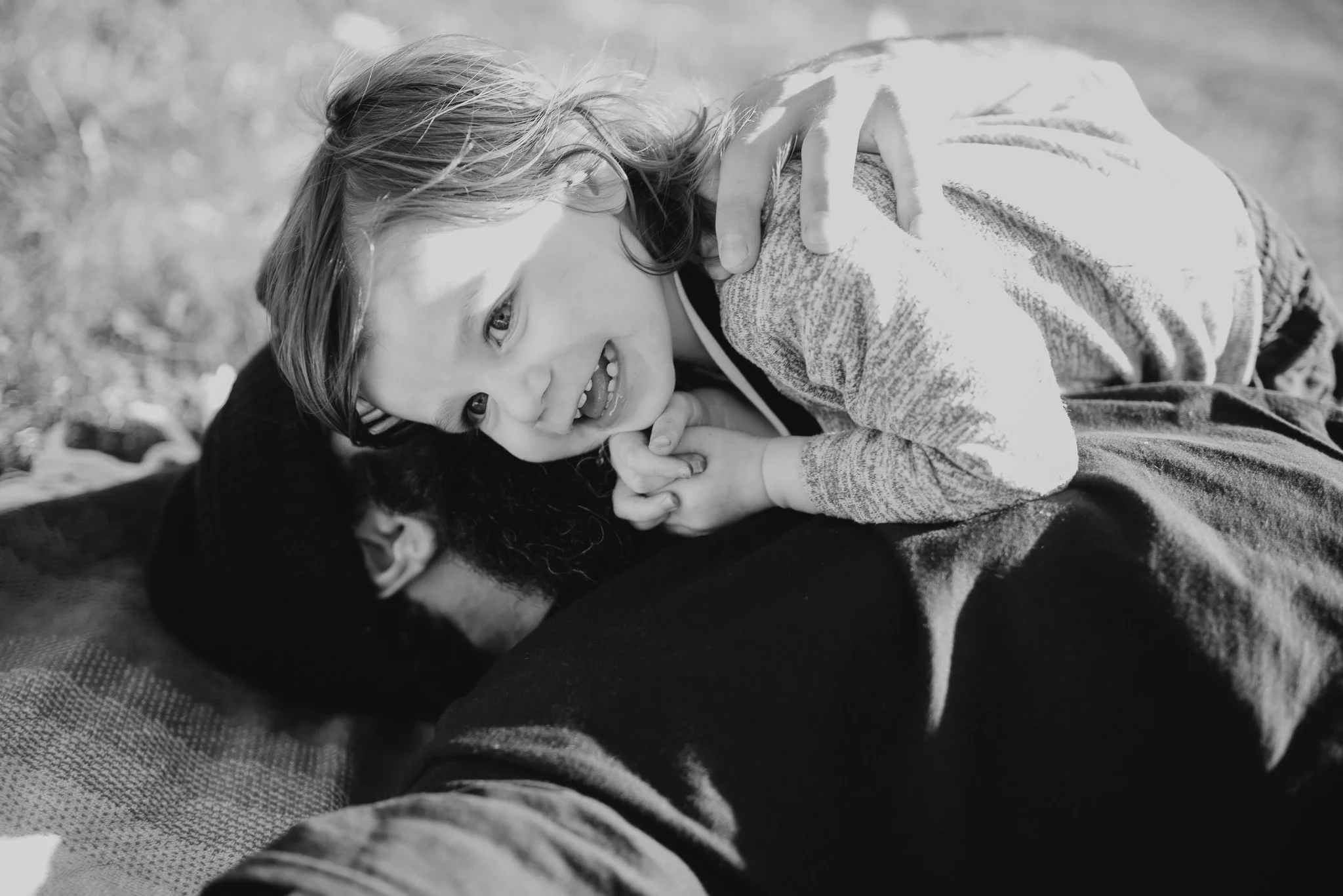 A young girl lying on her side on a blanket outdoors, smiling and looking at the camera.