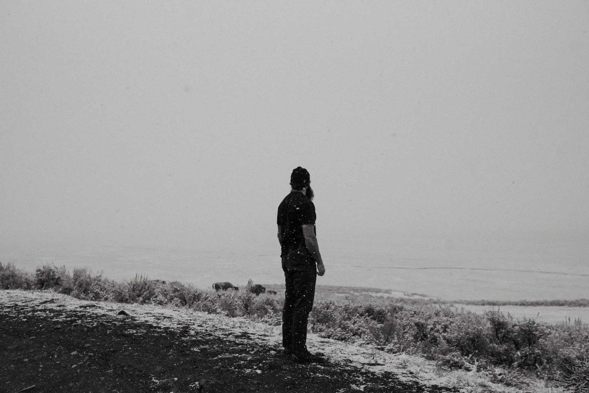 A man standing on a shoreline, looking out at the water, during snowy weather