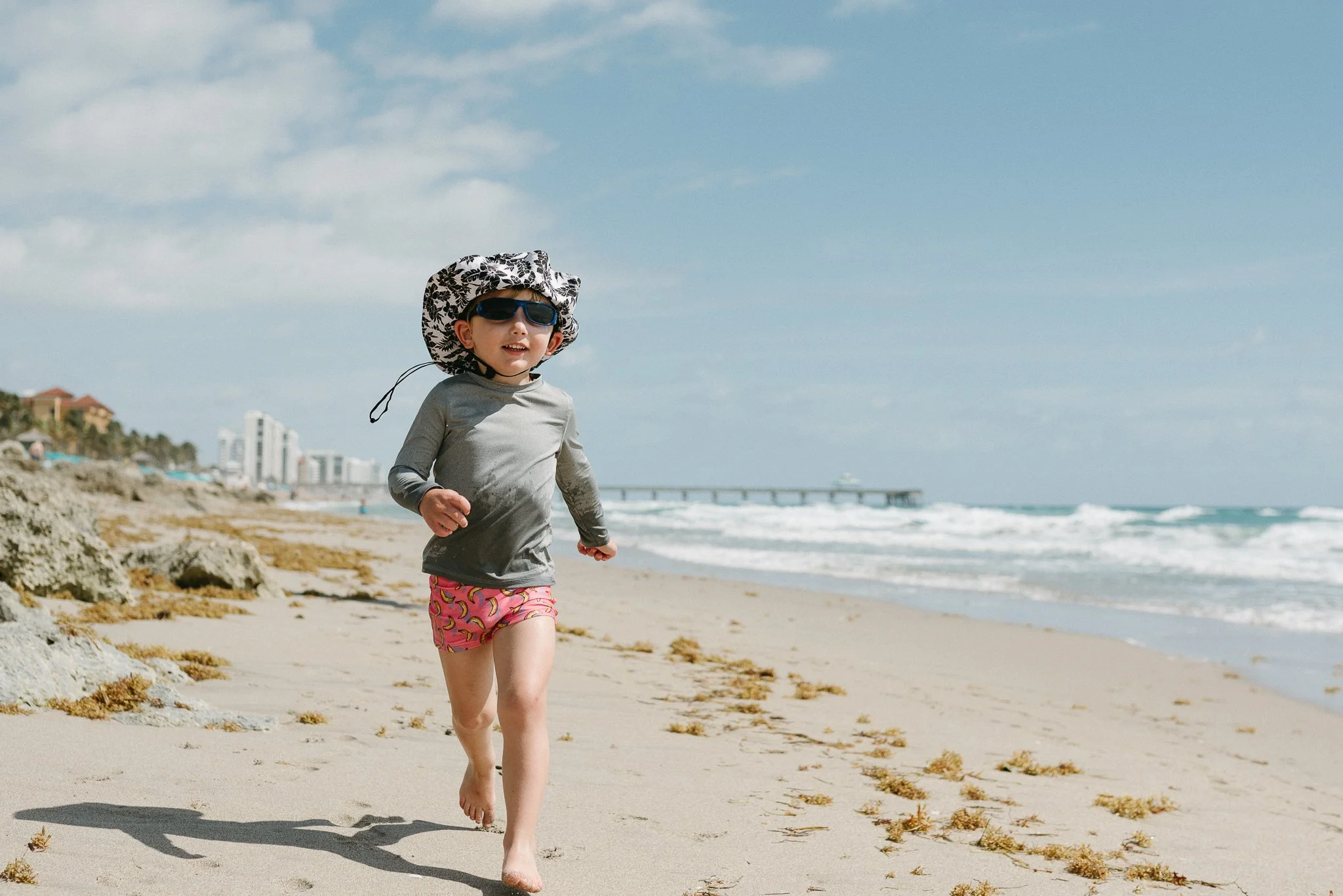 A young girl running barefoot on the beach, wearing a sun hat, sunglasses, a long-sleeve shirt, and pink shorts with bananas pattern, with the ocean, sky, and a pier in the background.