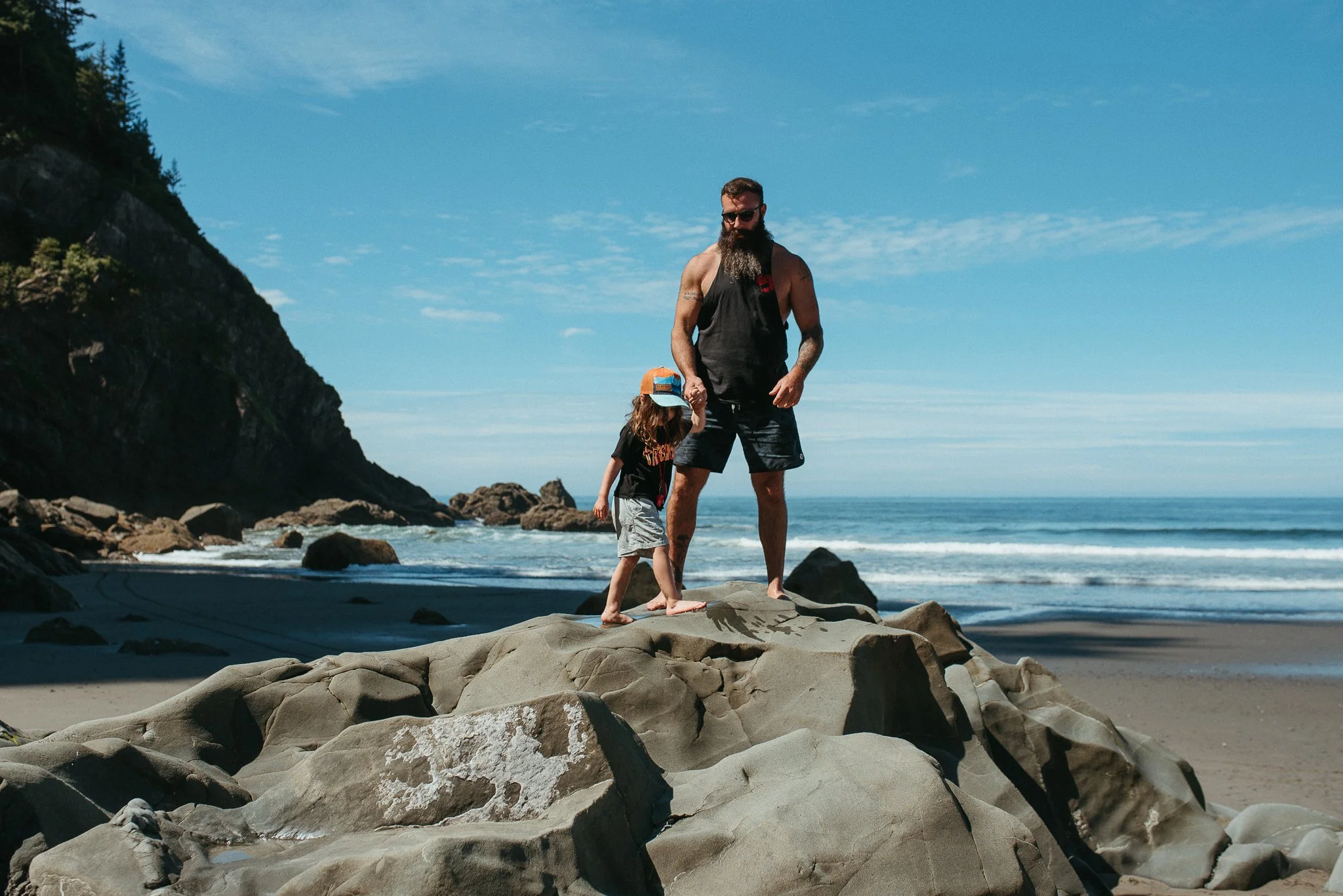 A bearded man and a young girl, standing on rocks at the beach, with the ocean and a cliff in the background on a clear day.