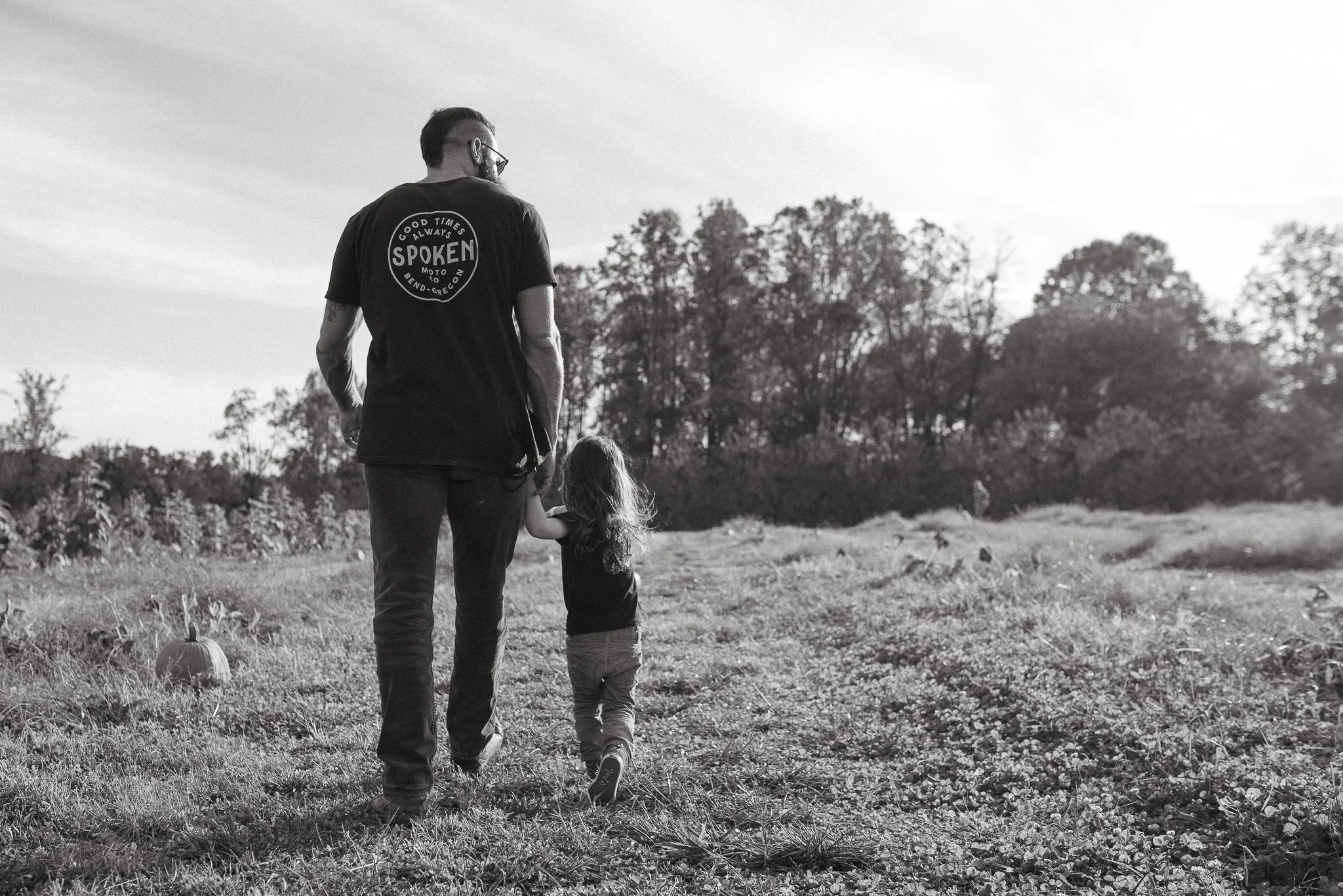 A man and a young girl walk hand in hand through a field with pumpkins, with trees in the background, during sunset or late afternoon, in black and white.