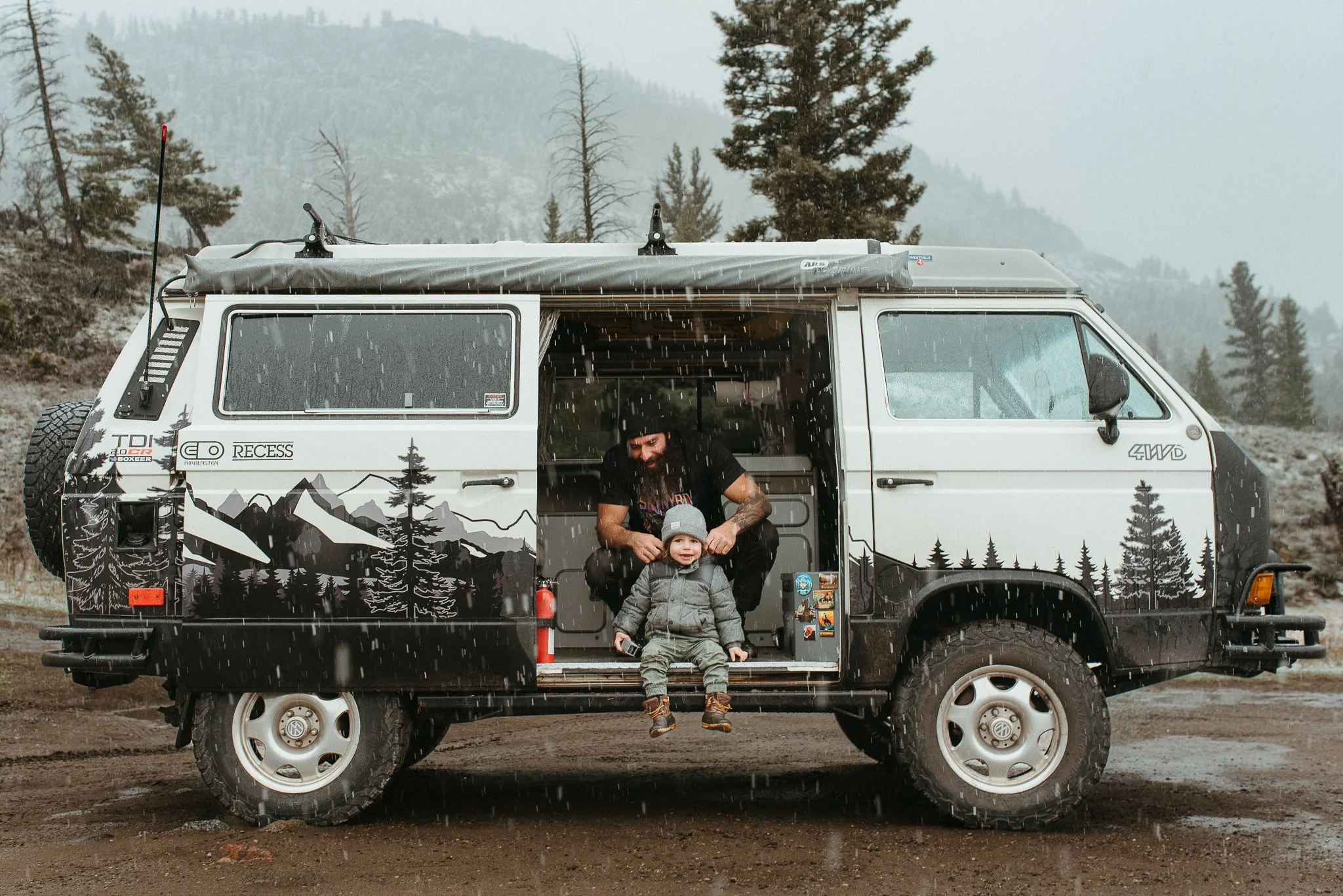 A camper van with mountain and forest artwork on its side is parked outdoors in rainy weather, with a bearded man and a young boy on the steps.