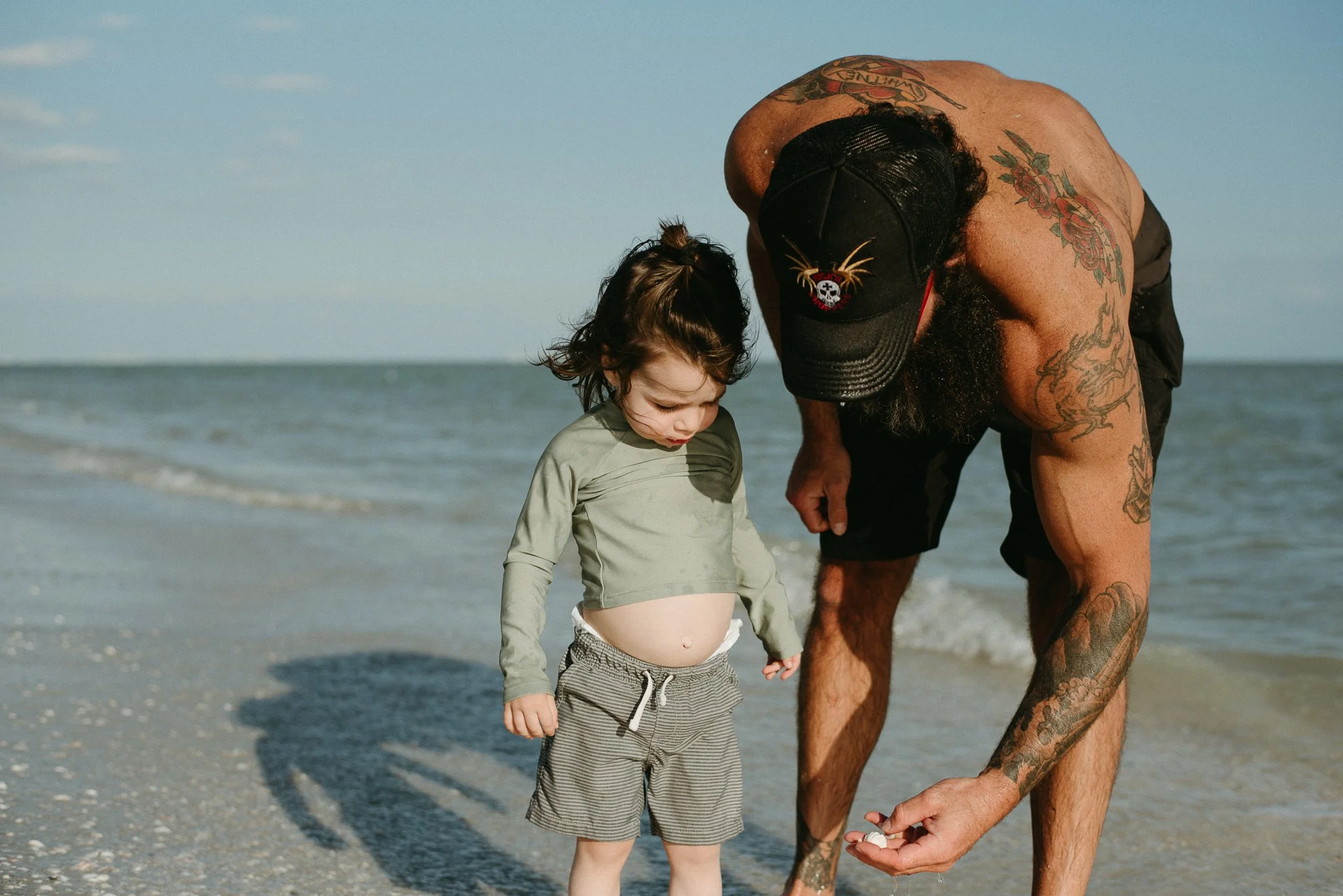 A young boy with a cloudy stomach standing on the beach next to an adult man with tattoos who is holding the boy's hand and looking down at him.