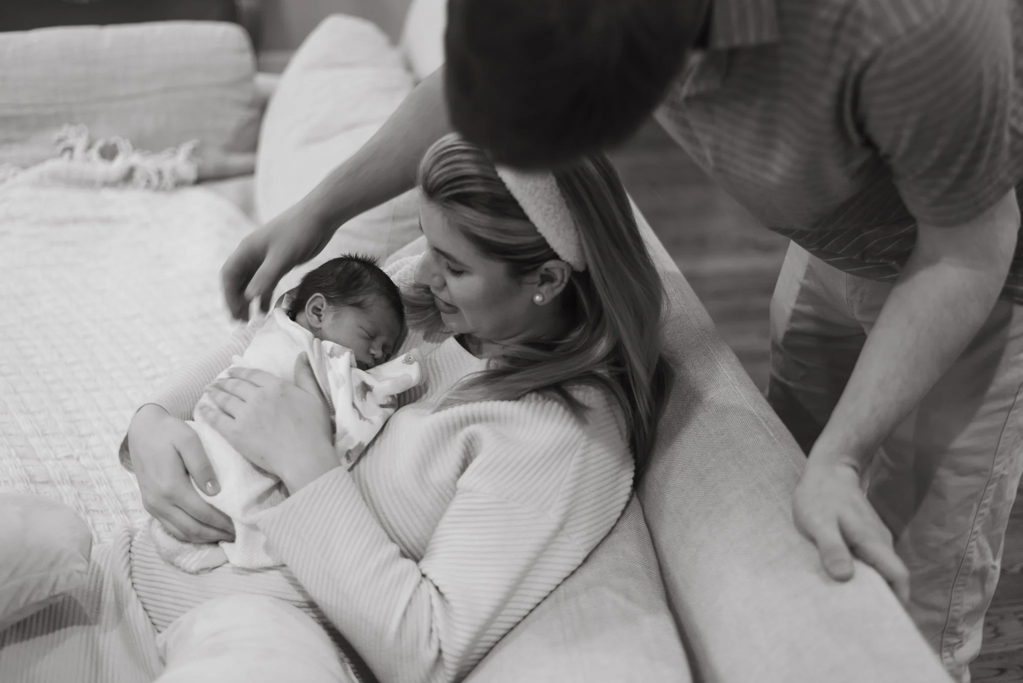 A woman holding a sleeping baby in her arms, lying on a bed, with a man leaning over them and gently touching the baby's head in a cozy, intimate setting.
