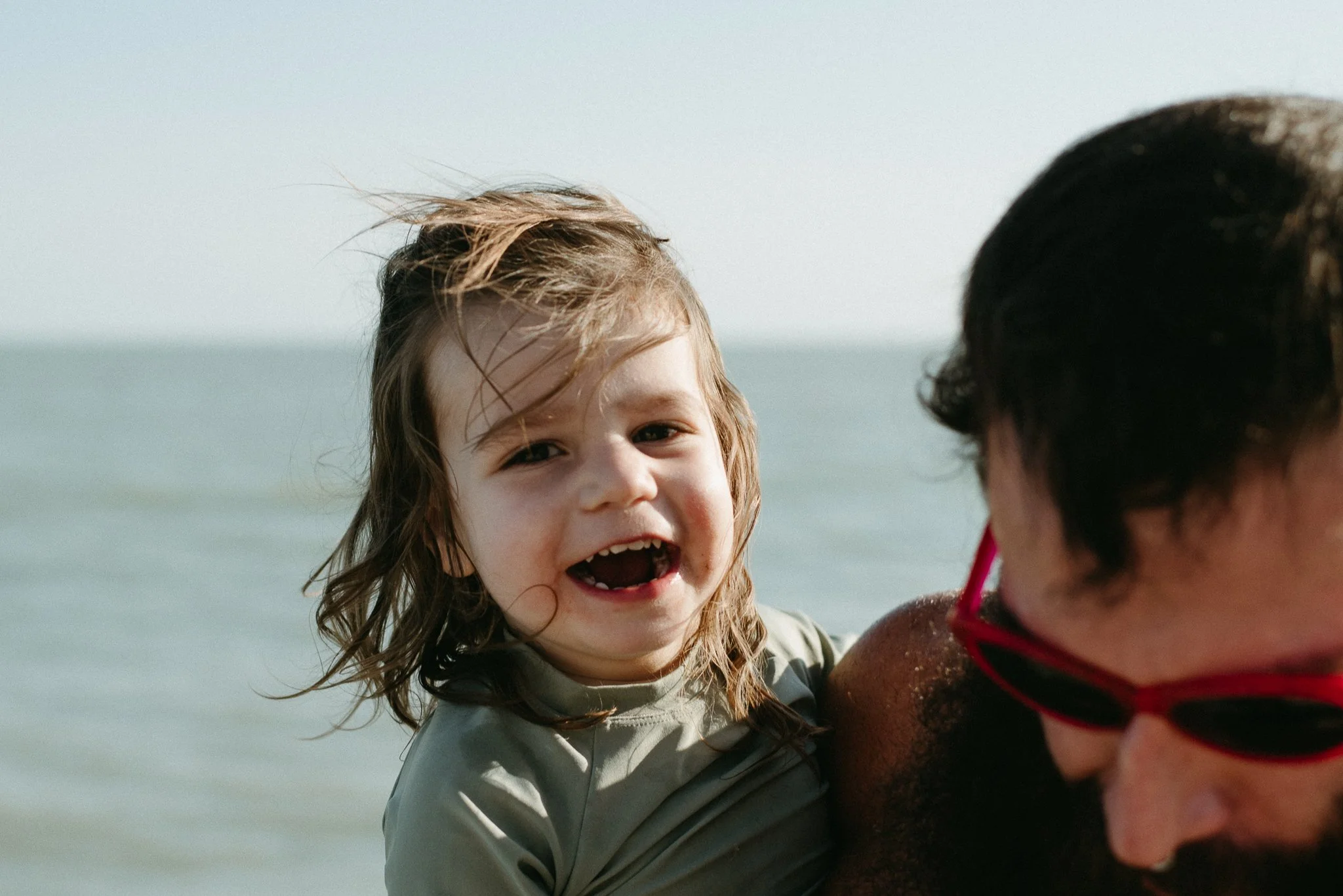 A smiling young girl with wet hair looking at a person wearing sunglasses and a swimsuit at the beach.