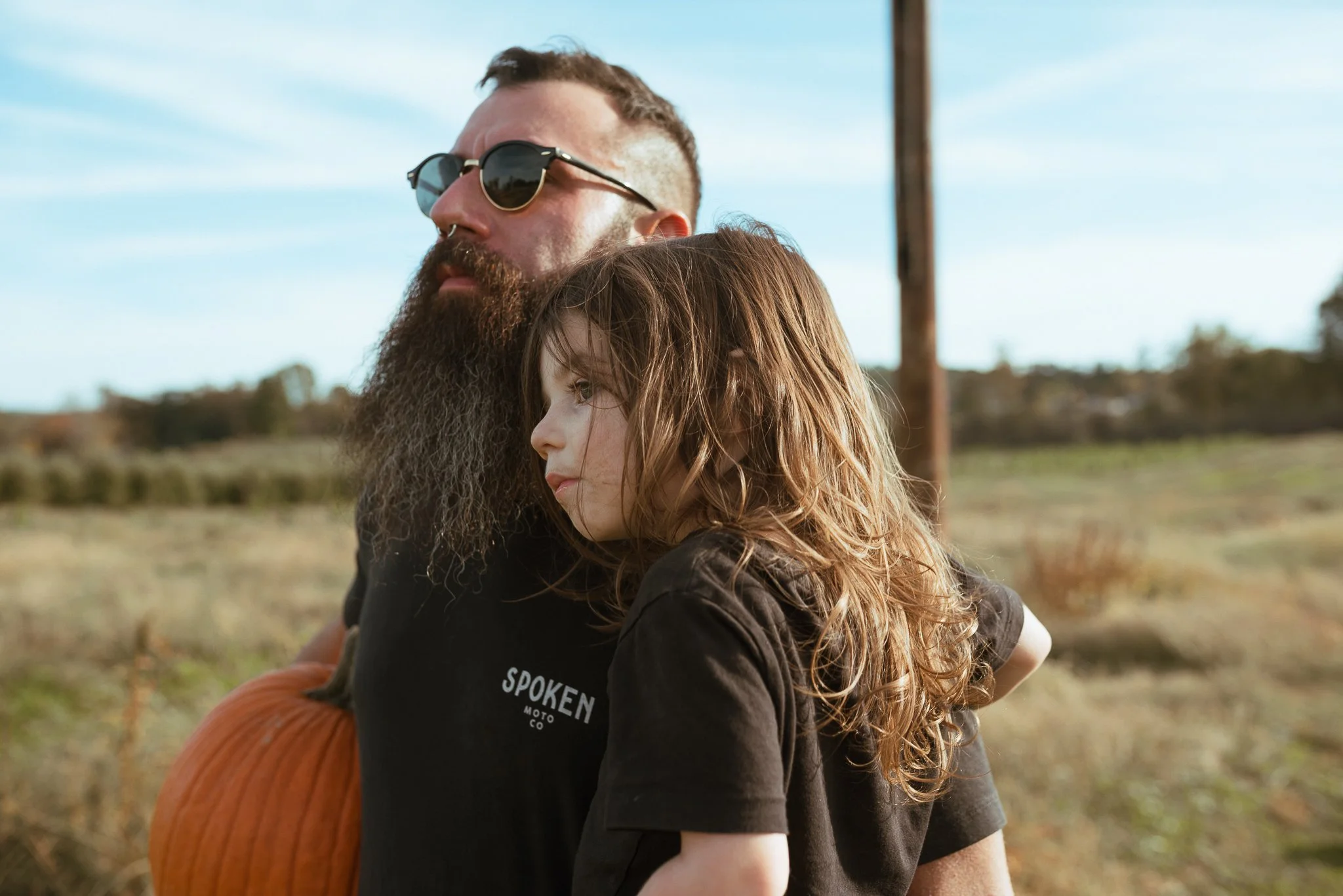 A bearded man wearing sunglasses and a black T-shirt with text, embracing a young girl with curly hair outdoors on a sunny day. The background features open fields with a utility pole and a blue sky with some clouds.