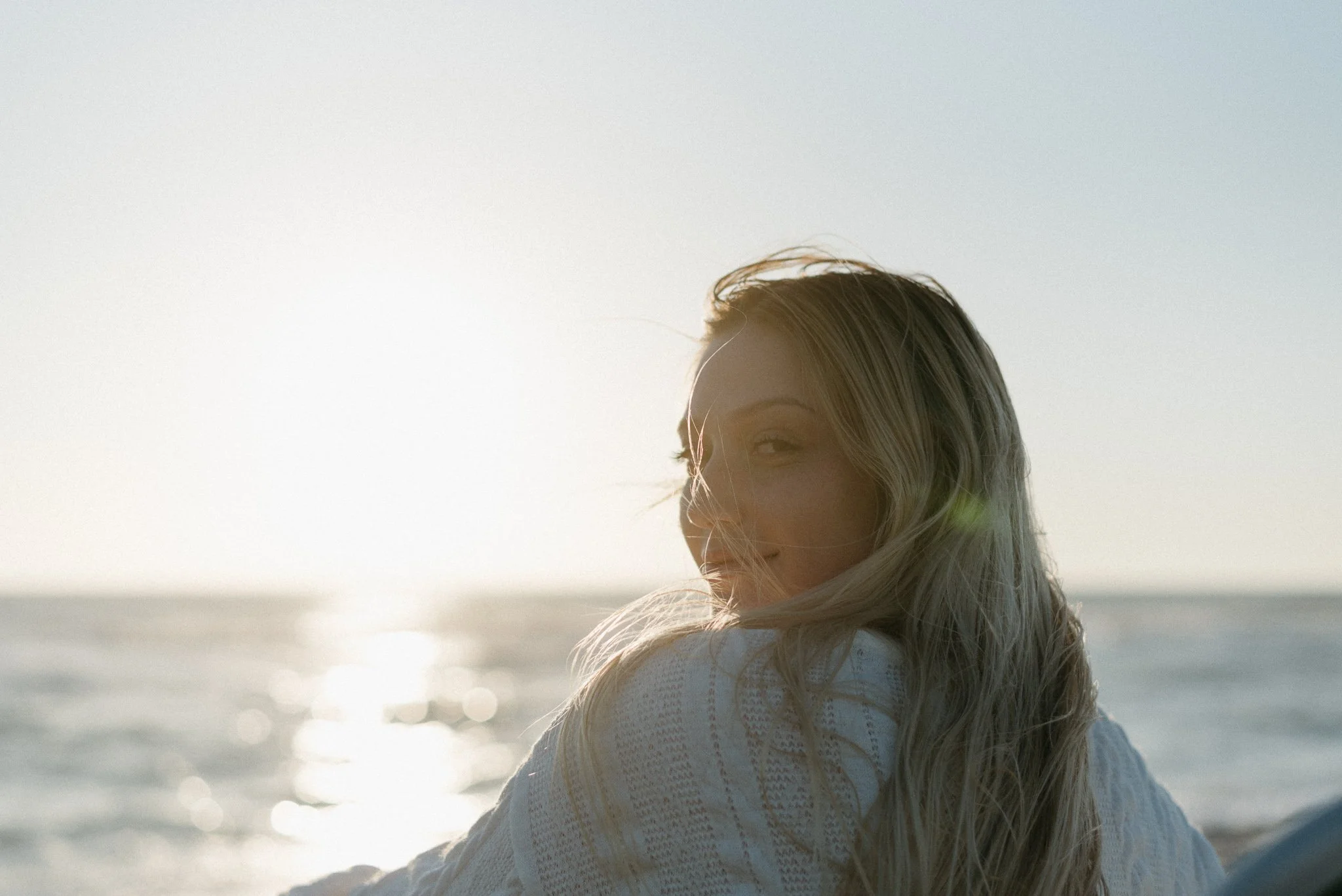 A woman with long blonde hair smiling at the beach during sunset.