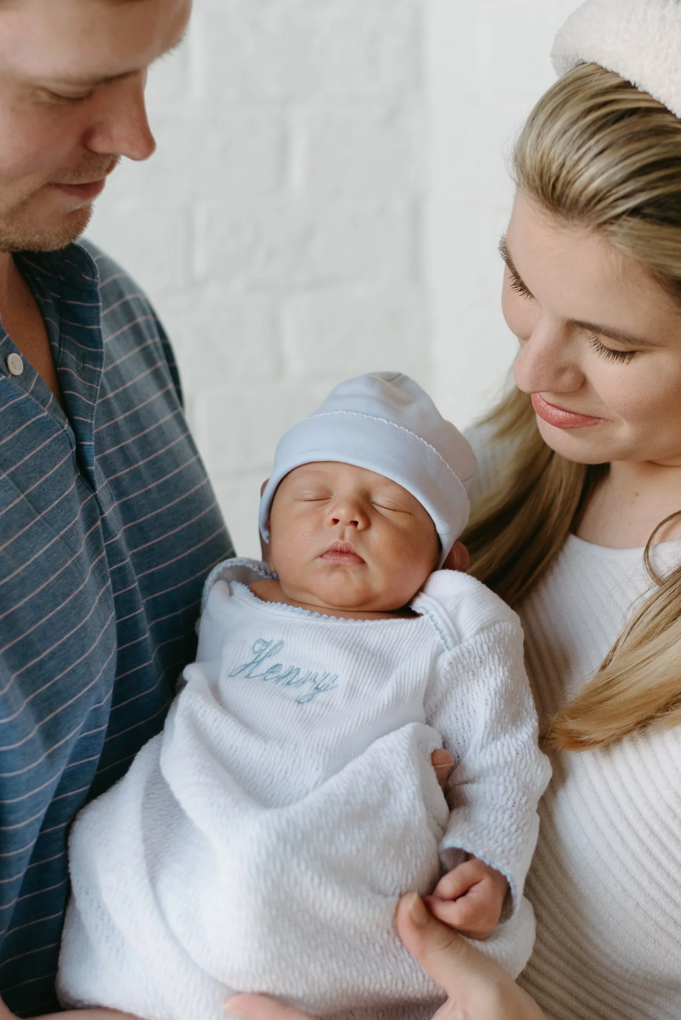 A family holds a sleeping baby dressed in white with embroidered text, with the mother and father looking at the baby affectionately.