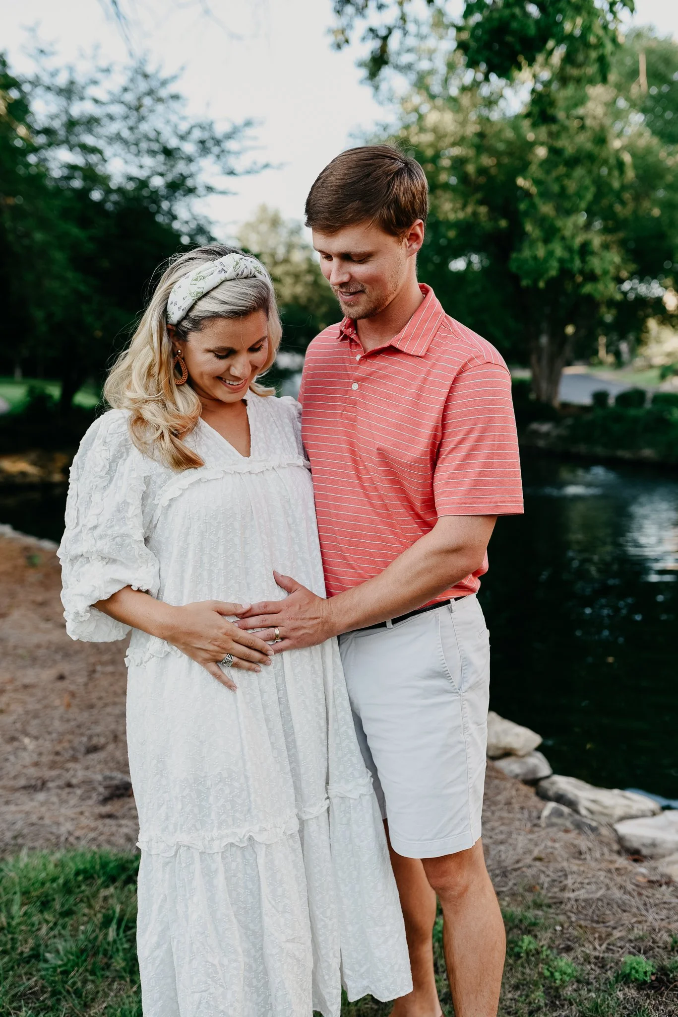A pregnant woman and a man standing outdoors near a body of water, smiling and gently holding her belly. The woman has blonde hair with a headband, and the man has brown hair, wearing a red striped shirt. They are surrounded by green trees and grass.