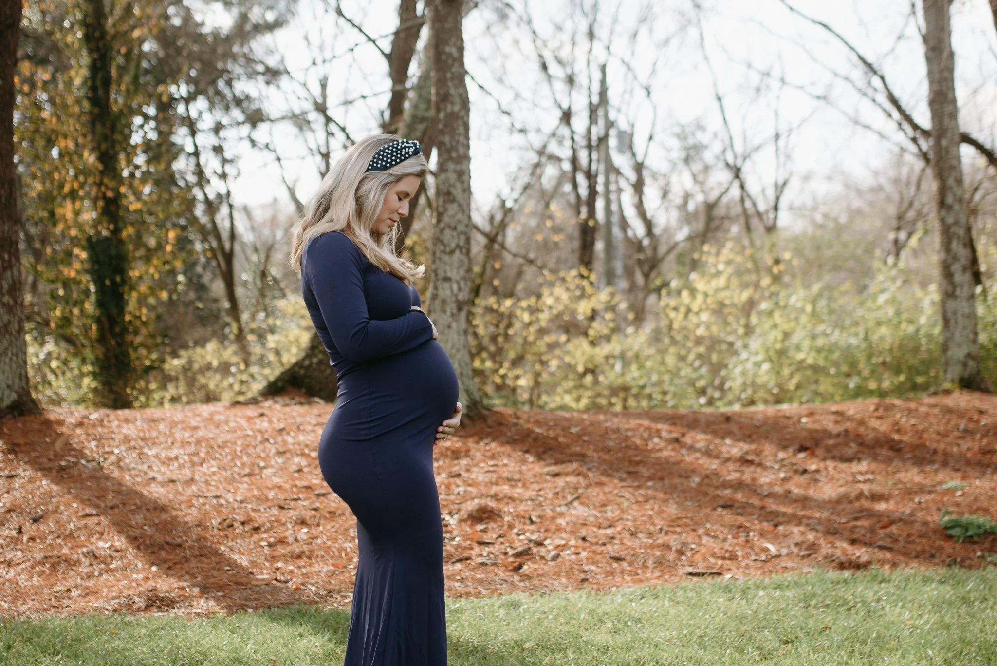 Pregnant woman in a blue dress standing outdoors among trees with fall foliage, holding her belly and looking down gently.