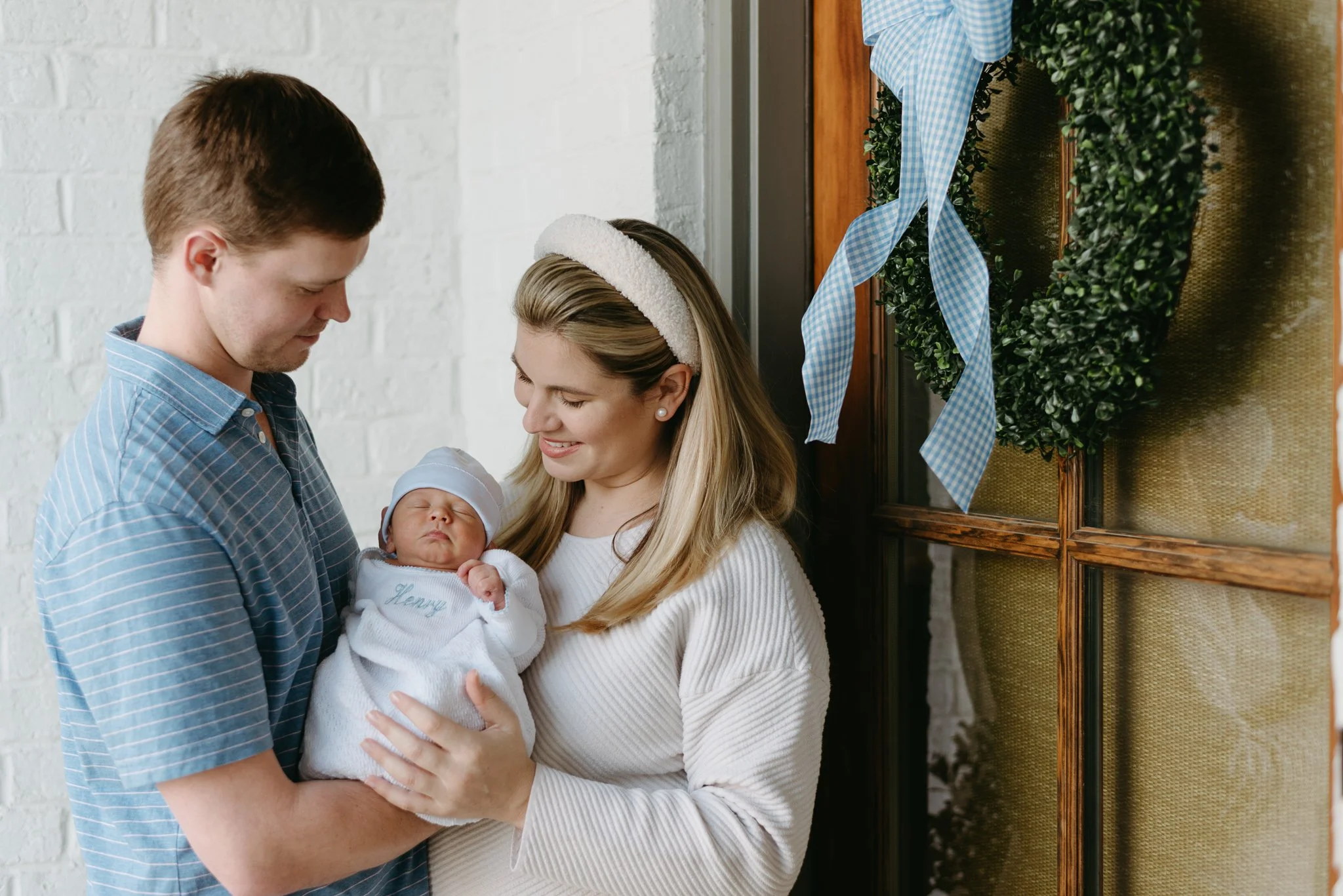 A woman holding a newborn baby, standing next to a man, in front of a decorated wall with a wreath and blue checkered ribbon.