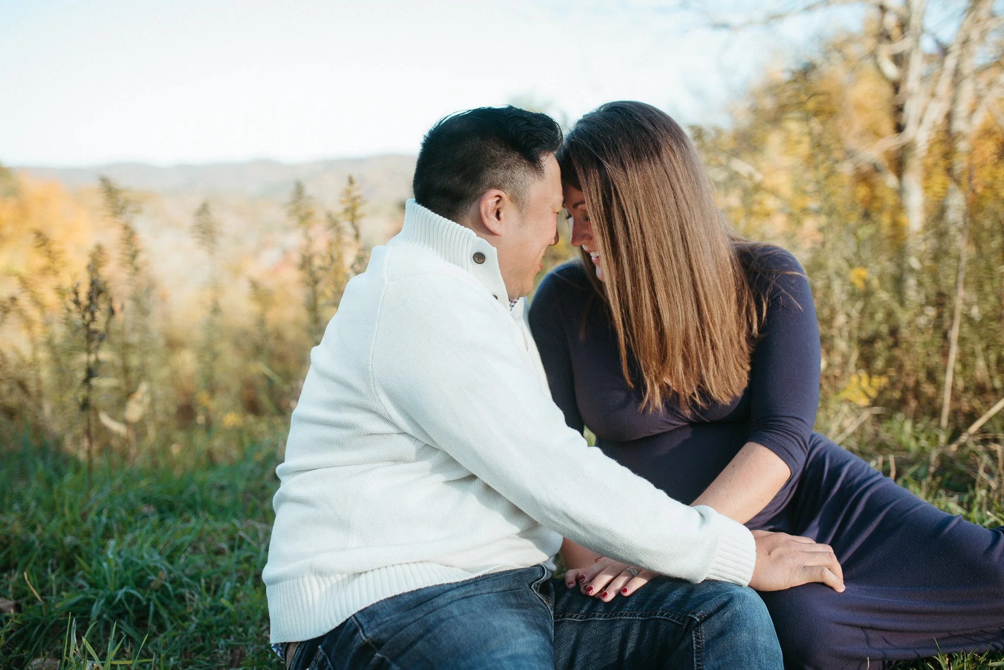 A happy couple is sitting close together outdoors in fall, with their foreheads touching and smiling, surrounded by trees with yellow leaves.