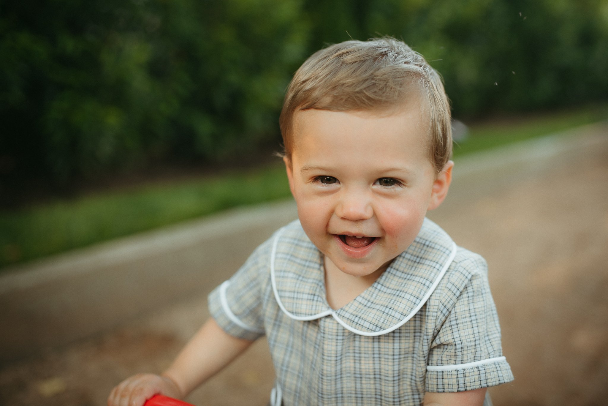 A young boy with light brown hair and blue eyes smiling outdoors, wearing a checkered shirt with white trim, in a park or natural setting.