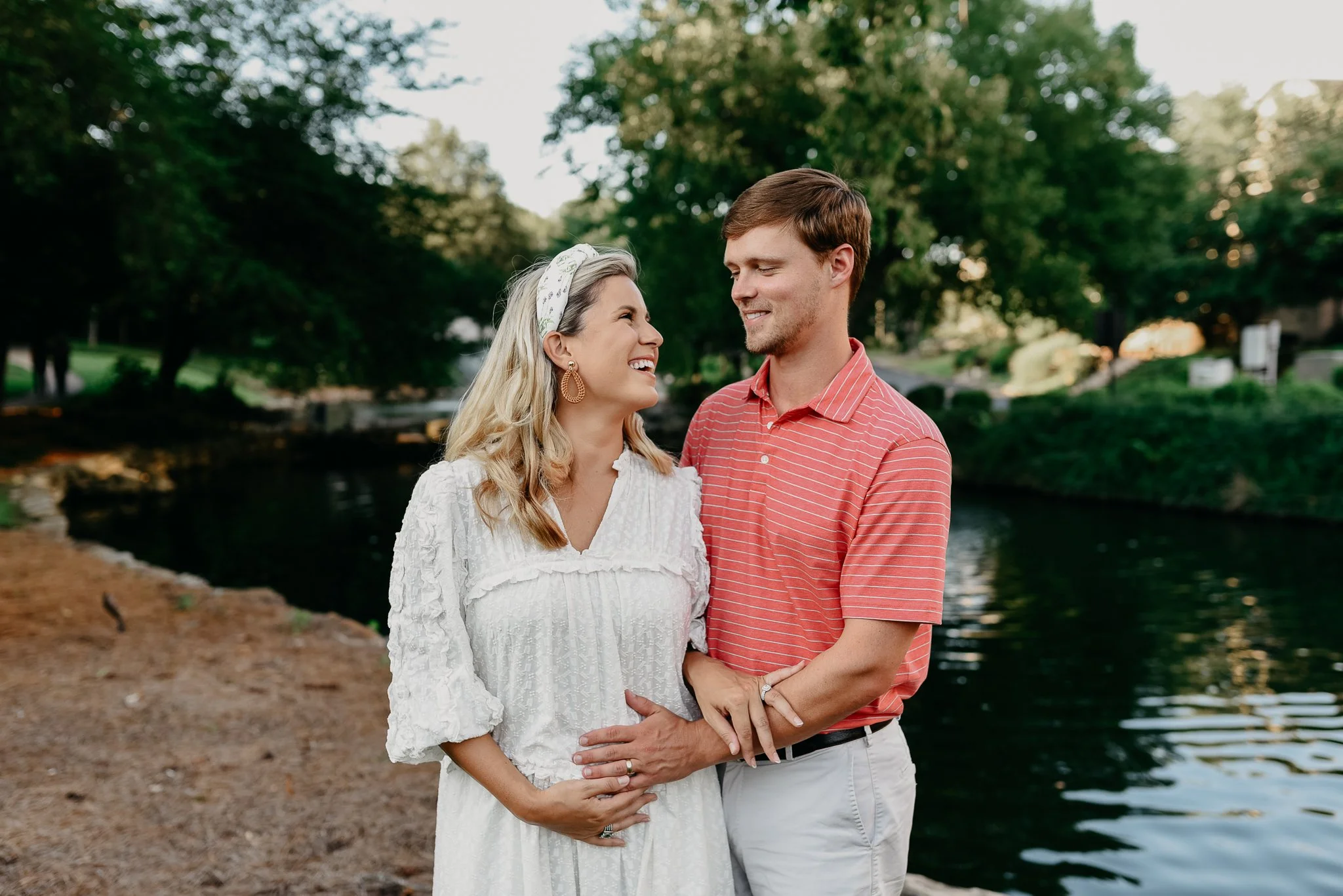 A smiling pregnant woman in a white dress and a man with a red striped shirt embrace by a river in a park with green trees.