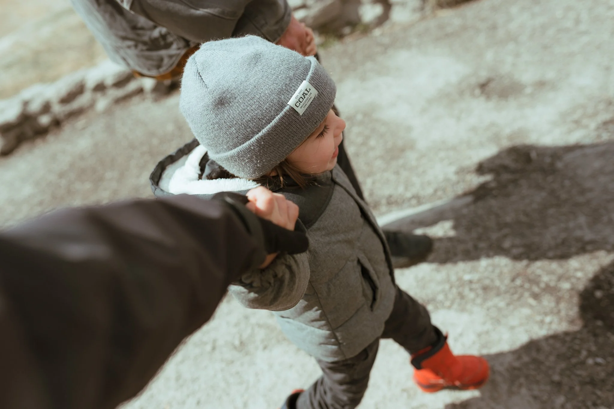 A young child wearing a gray beanie, gray jacket, and orange shoes, walking outdoors on a dirt path with an adult.
