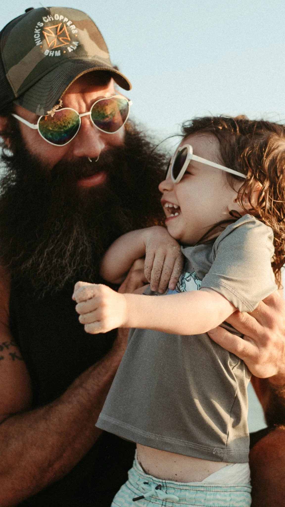 A man with a beard wearing sunglasses and a cap holding a young boy with curly hair and sunglasses, smiling.