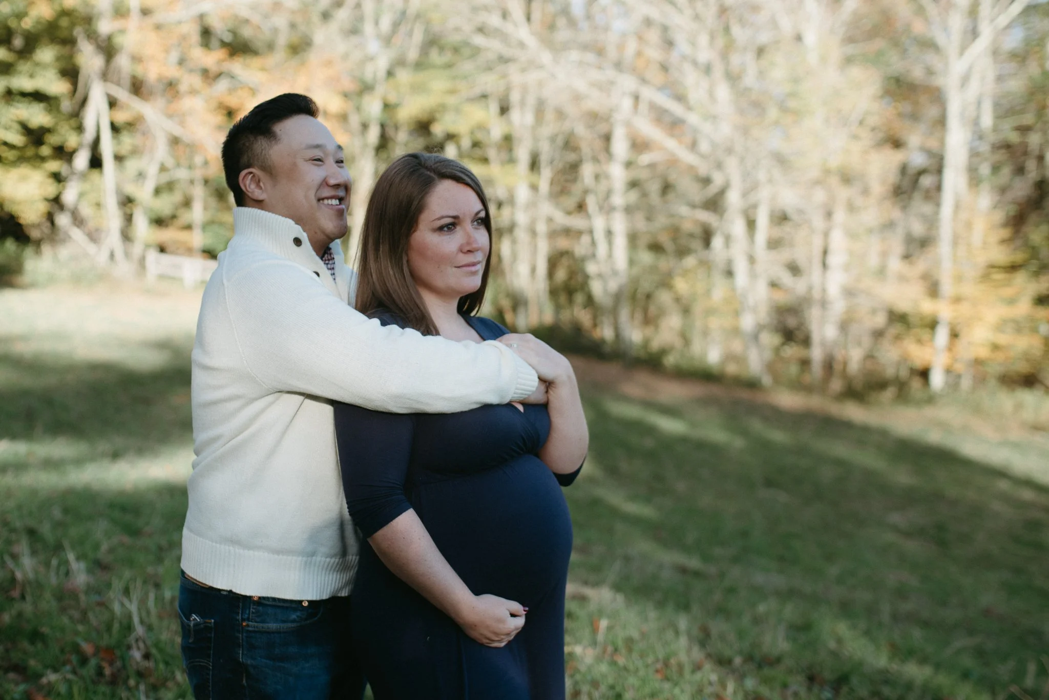 A couple, the man smiling and hugging a pregnant woman from behind in a park with trees in the background.
