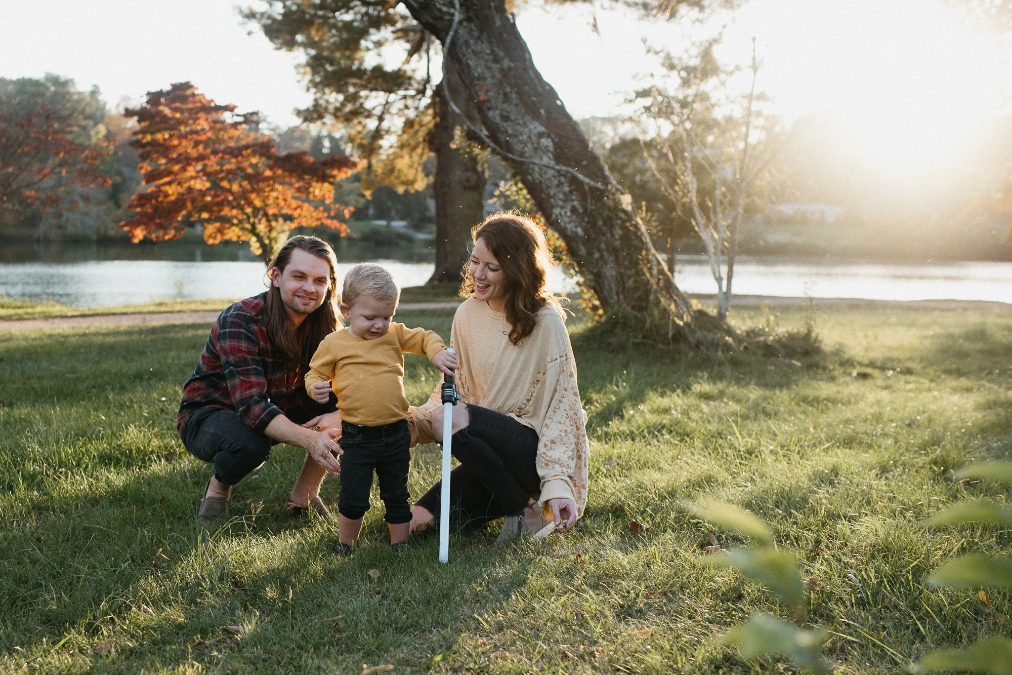 A family of three, including a man, woman, and a young child, sitting on the grass near a lake with trees showing autumn colors in the background during sunset.