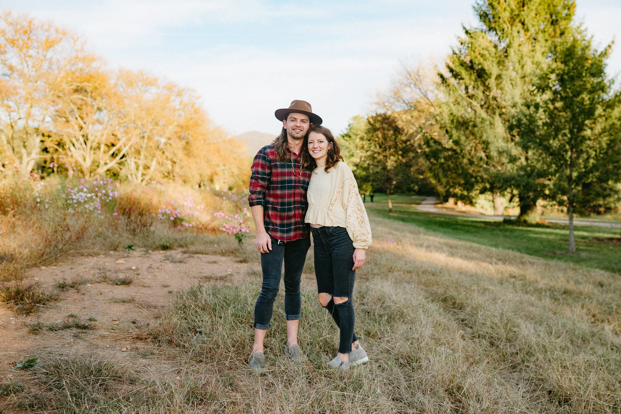 A young couple standing together outdoors in a park surrounded by trees and flowers during autumn, smiling at the camera.