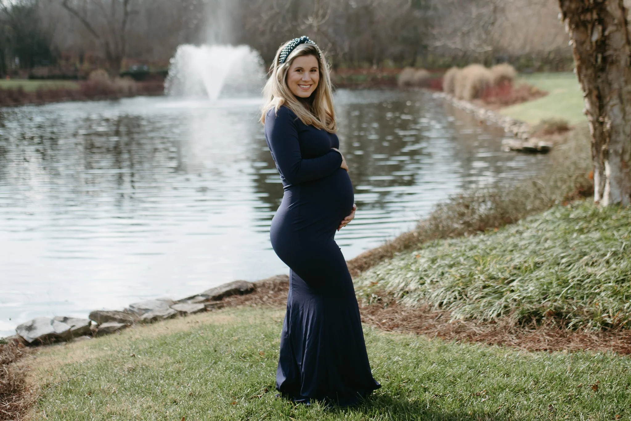 A pregnant woman wearing a navy blue dress and a pearl headband standing by a pond with a fountain, trees, and grass in the background.