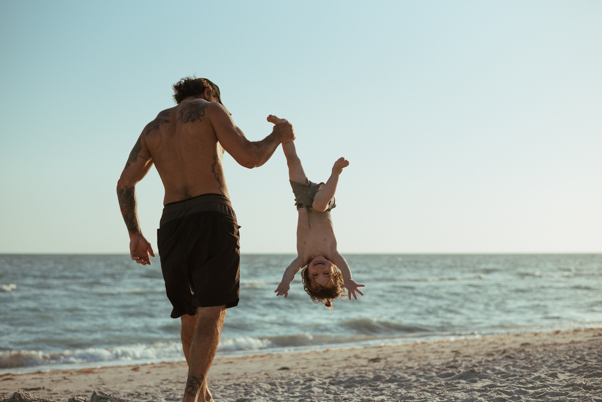 A shirtless man with tattoos holding a young girl upside down on a beach, both smiling with ocean and sky in the background.