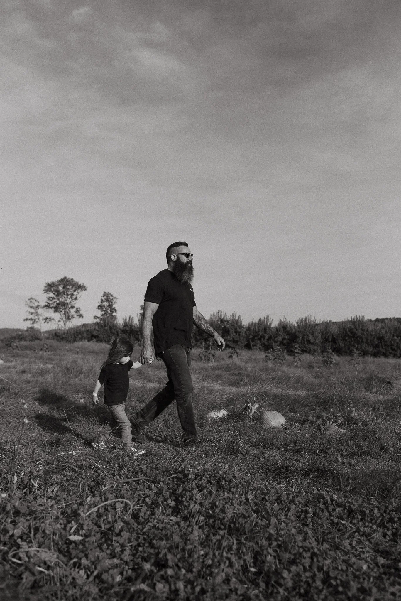 A man with a beard and sunglasses walks hand-in-hand with a young girl in a field of grass and small plants, with trees in the distance and a cloudy sky above, in a black and white photo.