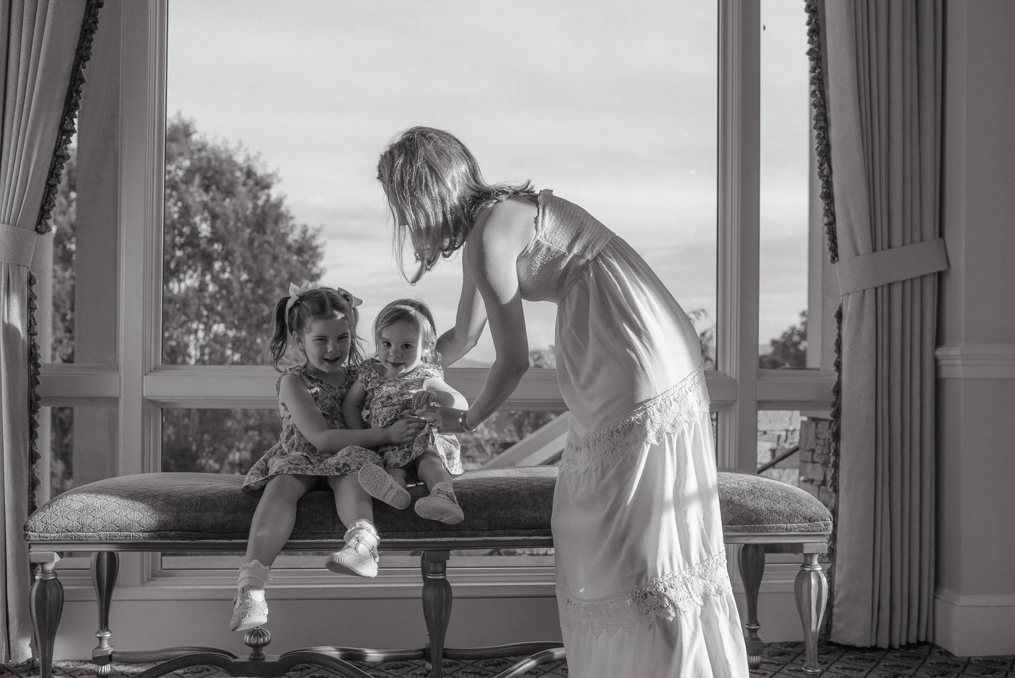 A woman with long hair and a sleeveless dress bends over to interact with two young girls sitting on a cushioned bench near a large open window. The girls, wearing dresses, are smiling and looking at the woman. Outside the window, trees and a cloudy 