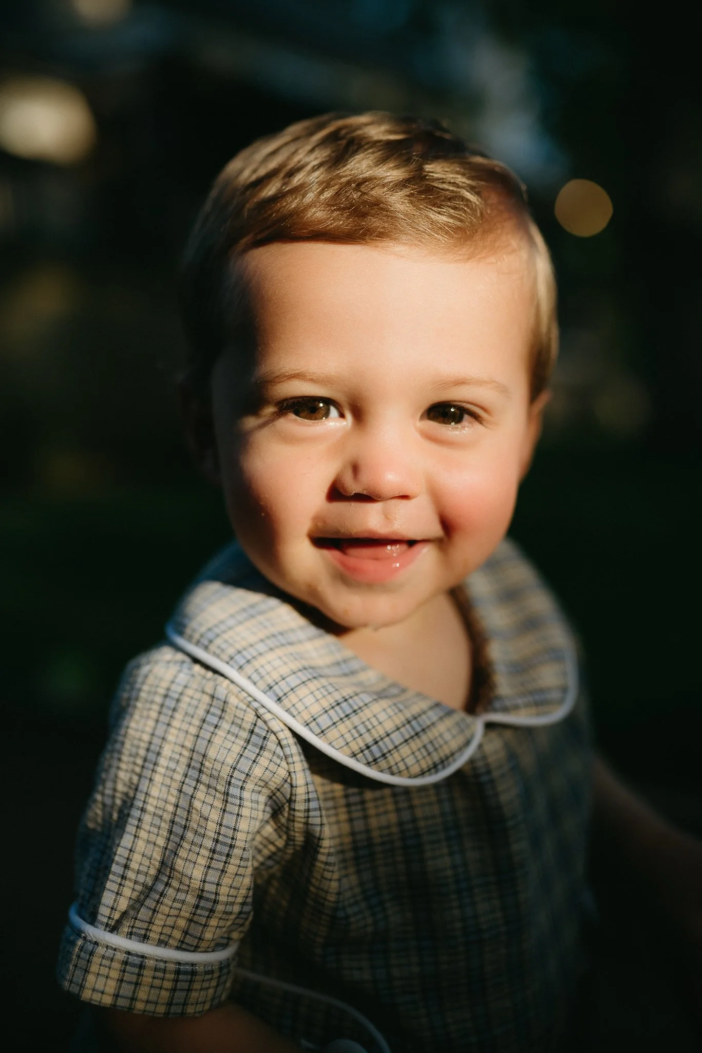 A young boy with light brown hair smiling outdoors at sunset, wearing a plaid shirt with a white piped collar.