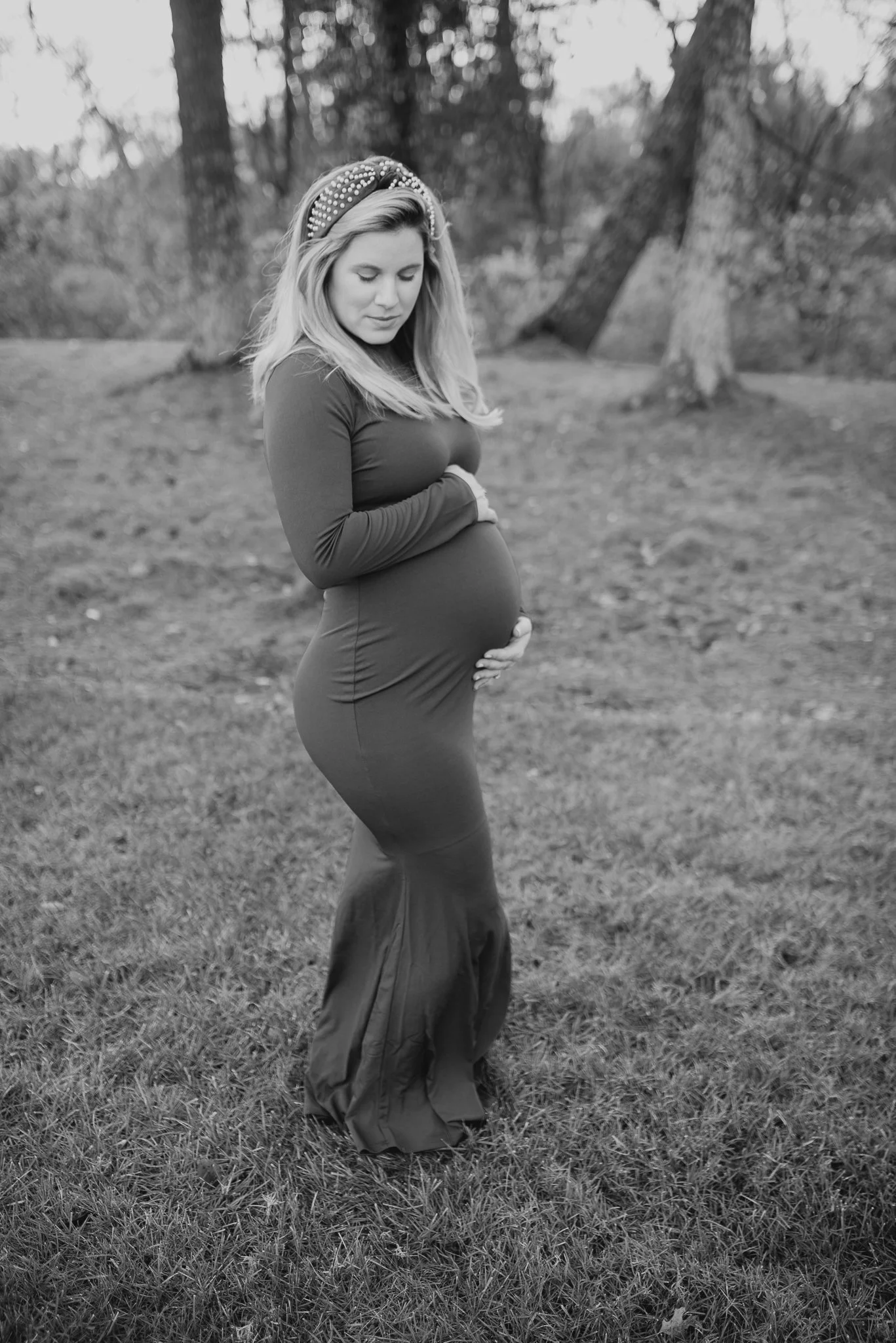 A pregnant woman wearing a headband with pearls, a long-sleeve top, and a flowing skirt is standing outdoors on grass, gently holding her belly and looking down with a serene expression. The background features trees and a natural setting.